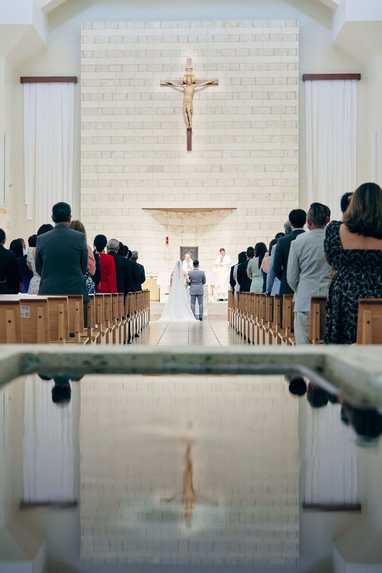 Wedding ceremony in a modern Catholic church with guests standing as couple exchanges vows at the altar.
