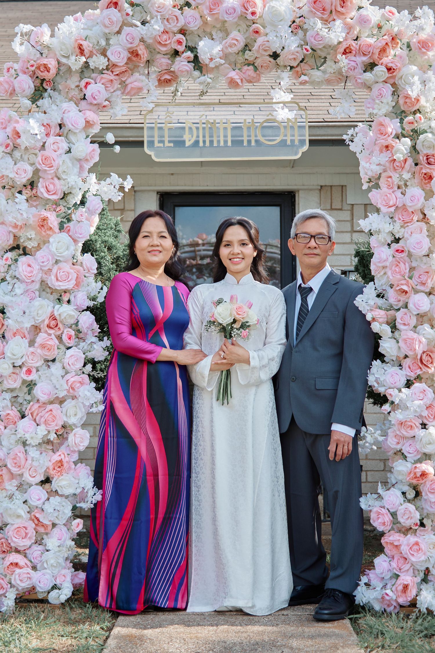Bride in white ao dai holds bouquet flanked by parents under floral arch of pink and white roses at Vietnamese wedding.