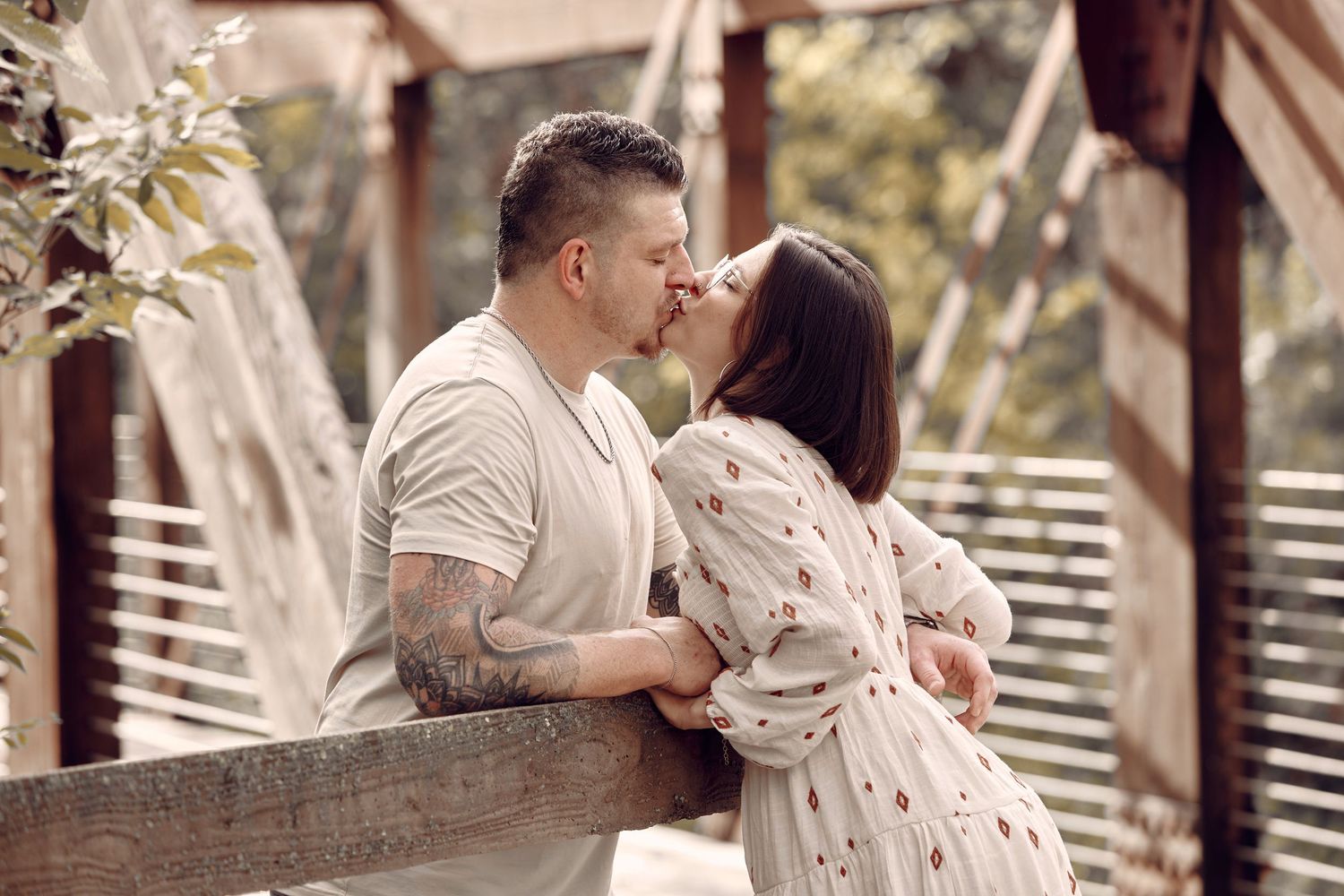 Couple sharing a tender kiss on a wooden bridge surrounded by greenery.