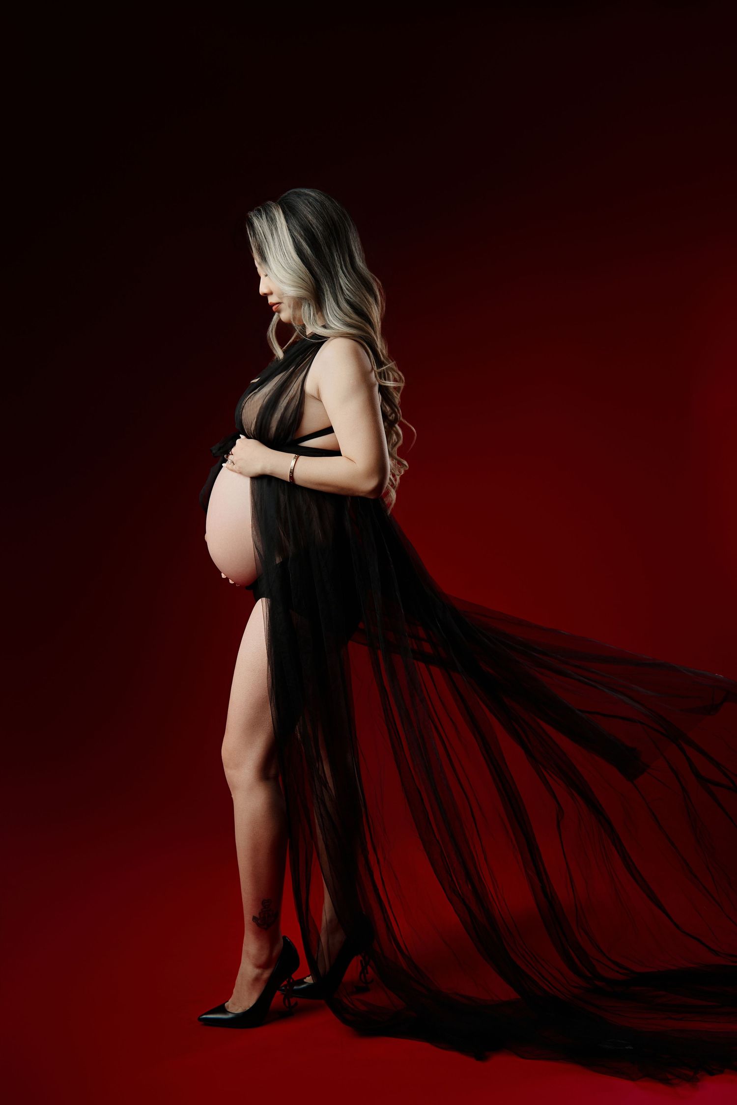 Pregnant woman in flowing black sheer gown with train posing on dark red background.