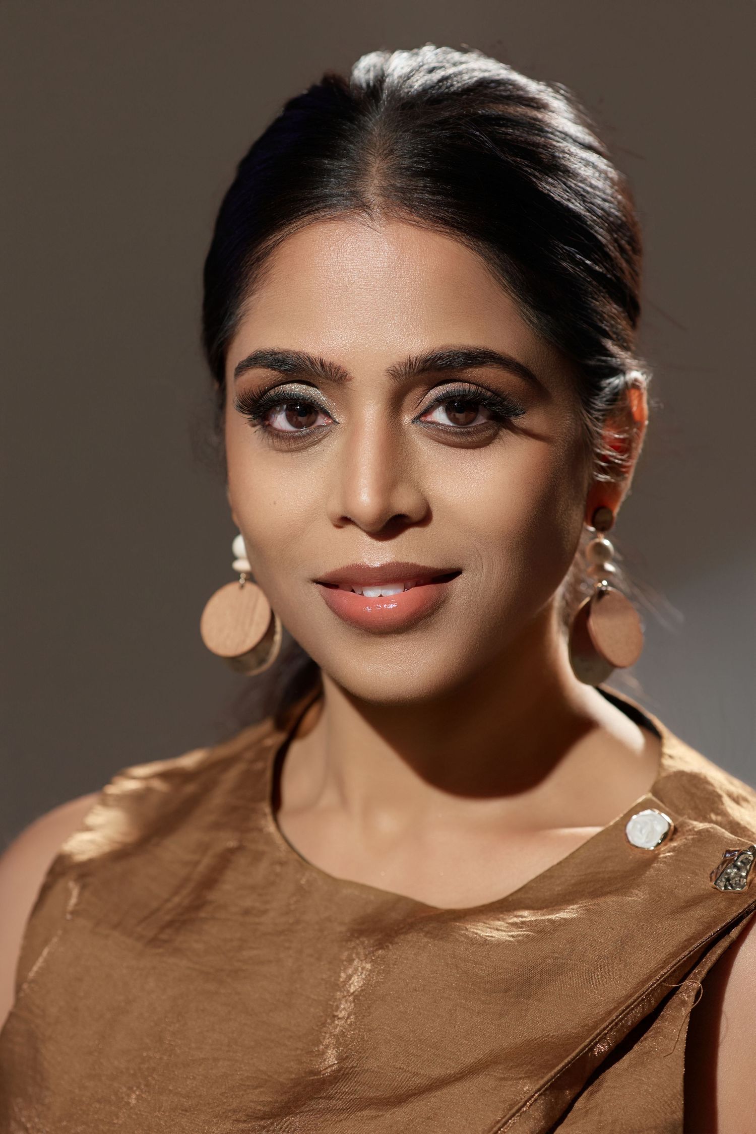 Close-up portrait of a young Indian woman with hoop earrings wearing a brown outfit in warm lighting.