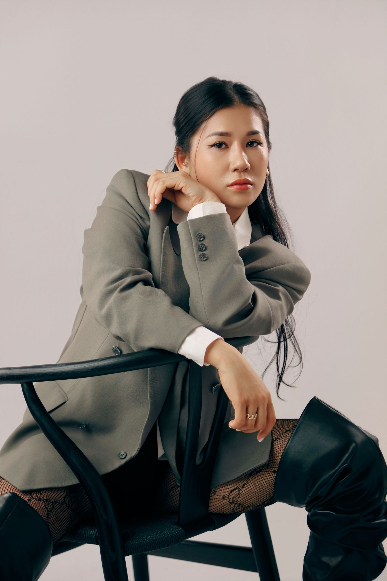Stylish Asian woman in oversized gray blazer poses on black chair with fishnet stockings.