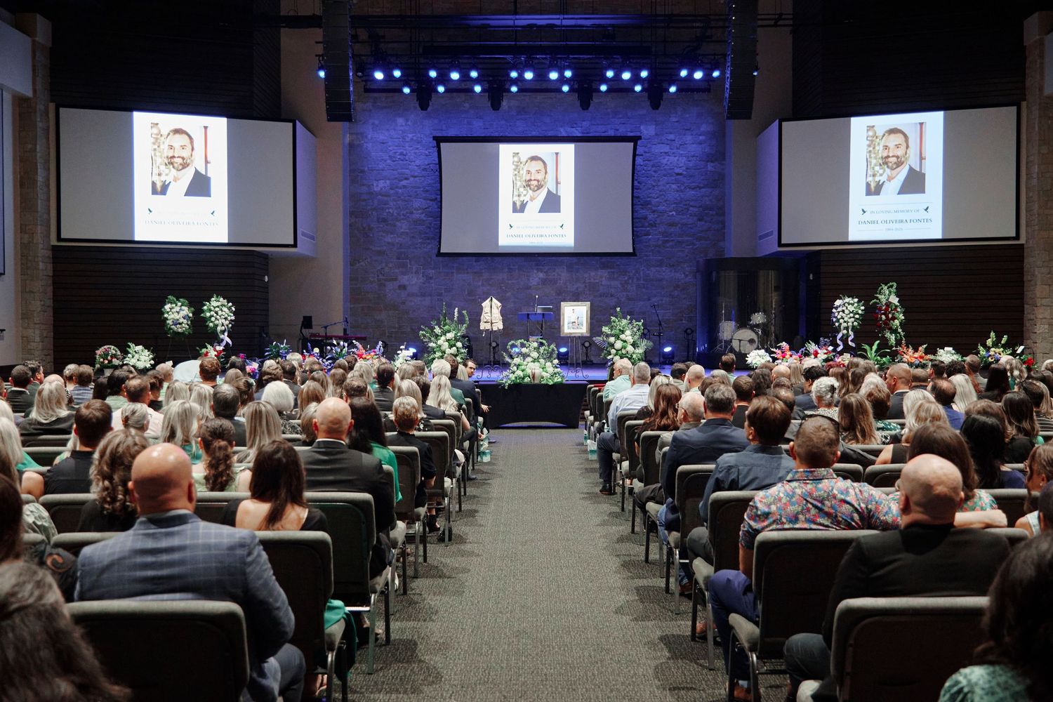 Large memorial service with hundreds of attendees seated in an auditorium with portrait displayed on screens.