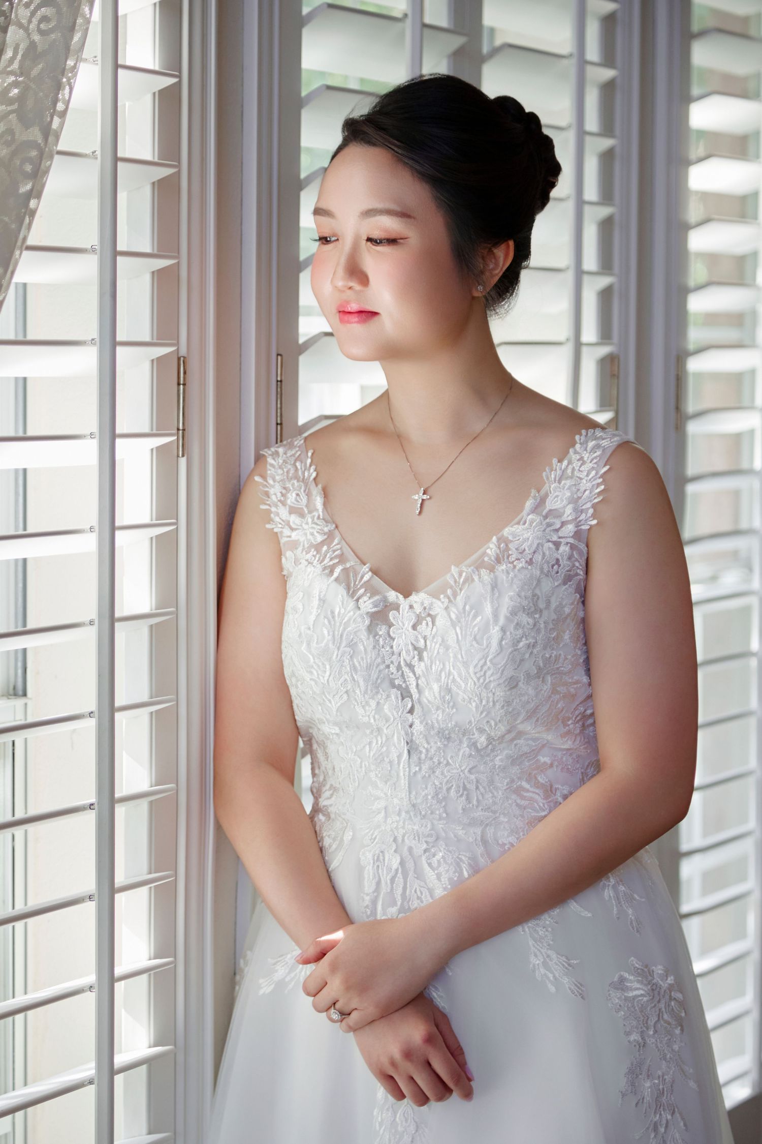 Asian bride in lace wedding gown standing by window with soft natural light, wearing cross necklace.