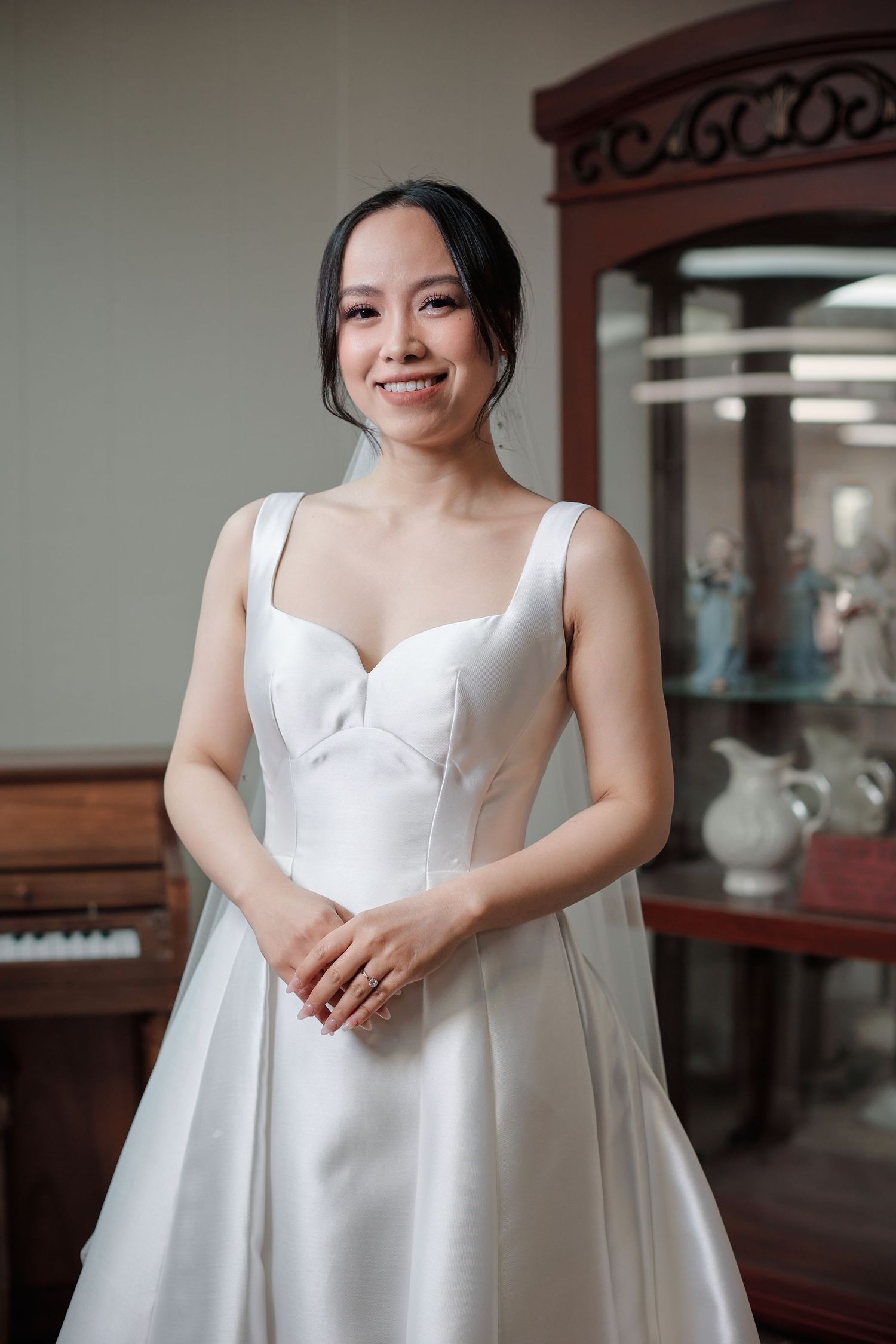 Beautiful Asian bride smiling in stunning white satin A-line wedding dress indoors.