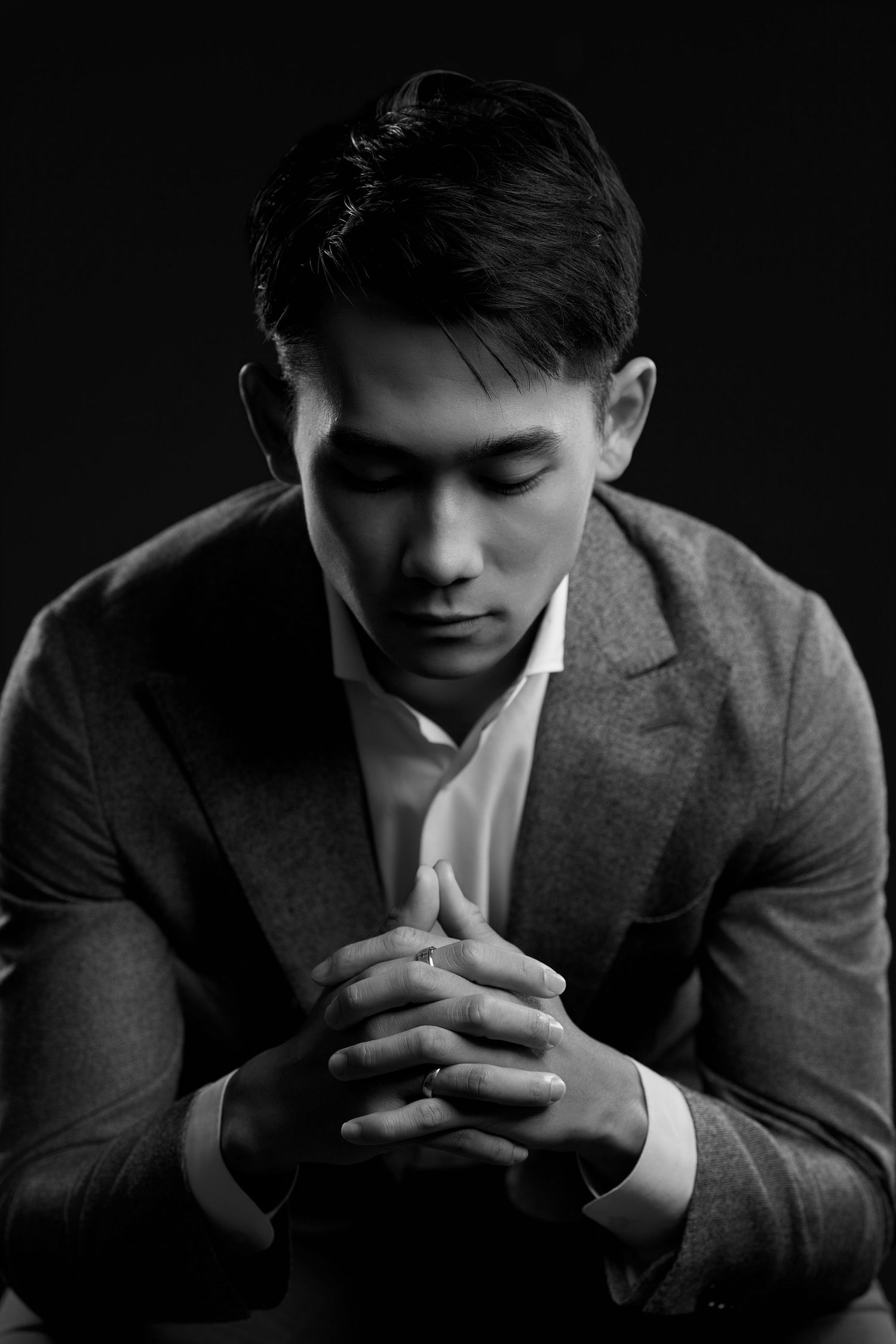 Black and white portrait of a young Asian man in a suit with clasped hands, eyes downcast, against dark background.