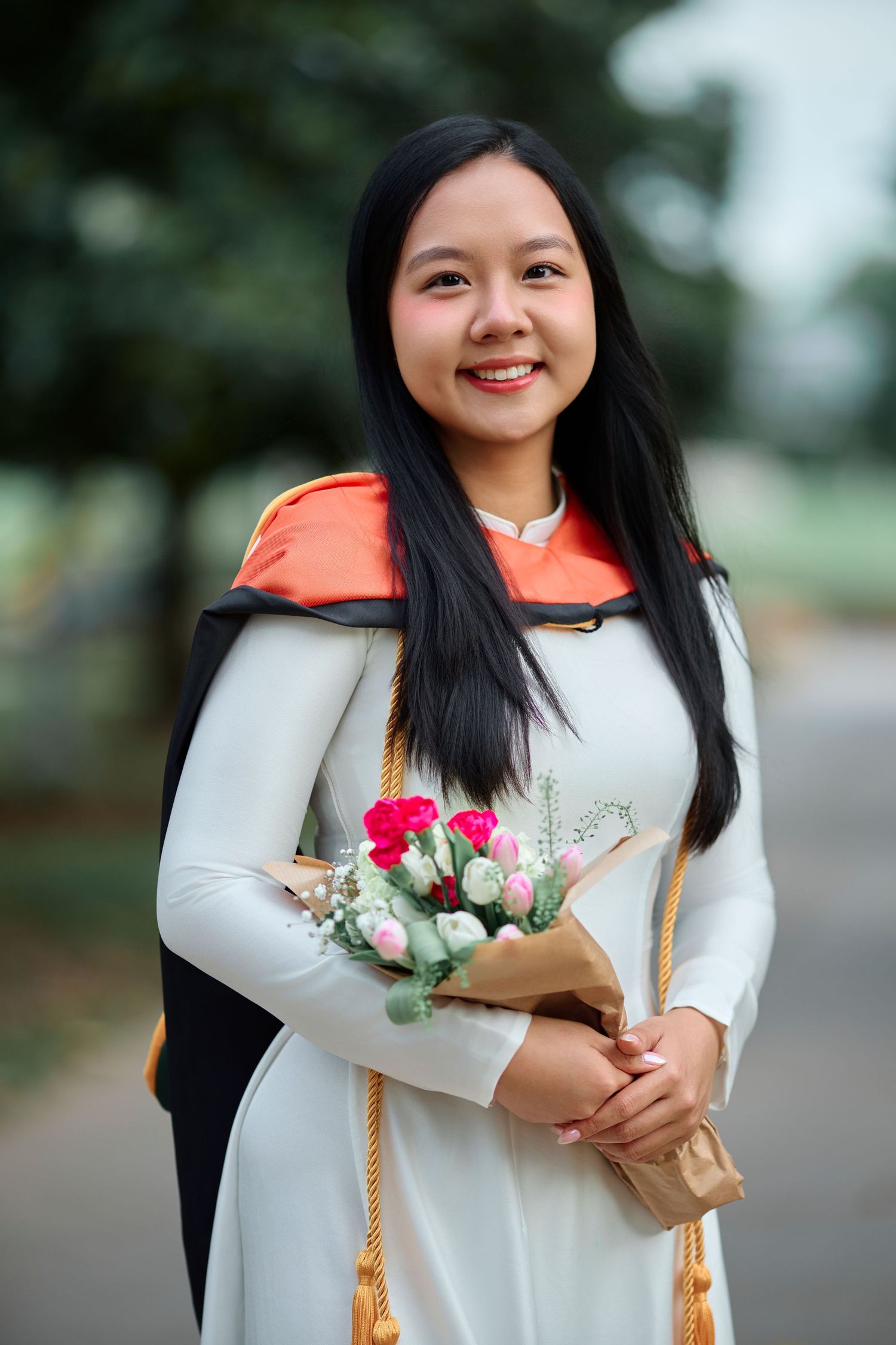 Smiling young woman in white ao dai and graduation regalia holding a bouquet of pink roses outdoors.