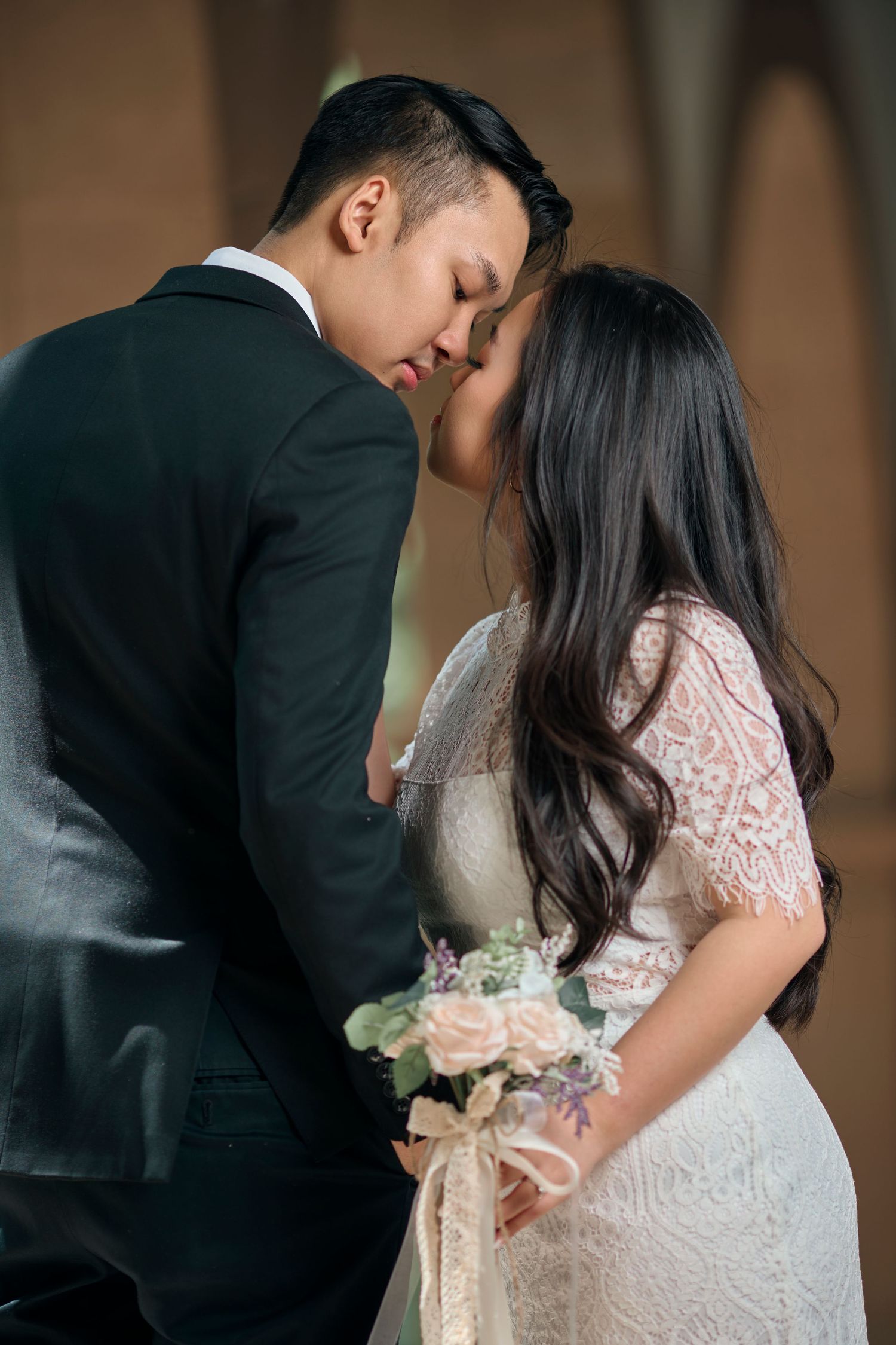 Asian bride and groom sharing tender forehead touch moment with wedding bouquet indoors.