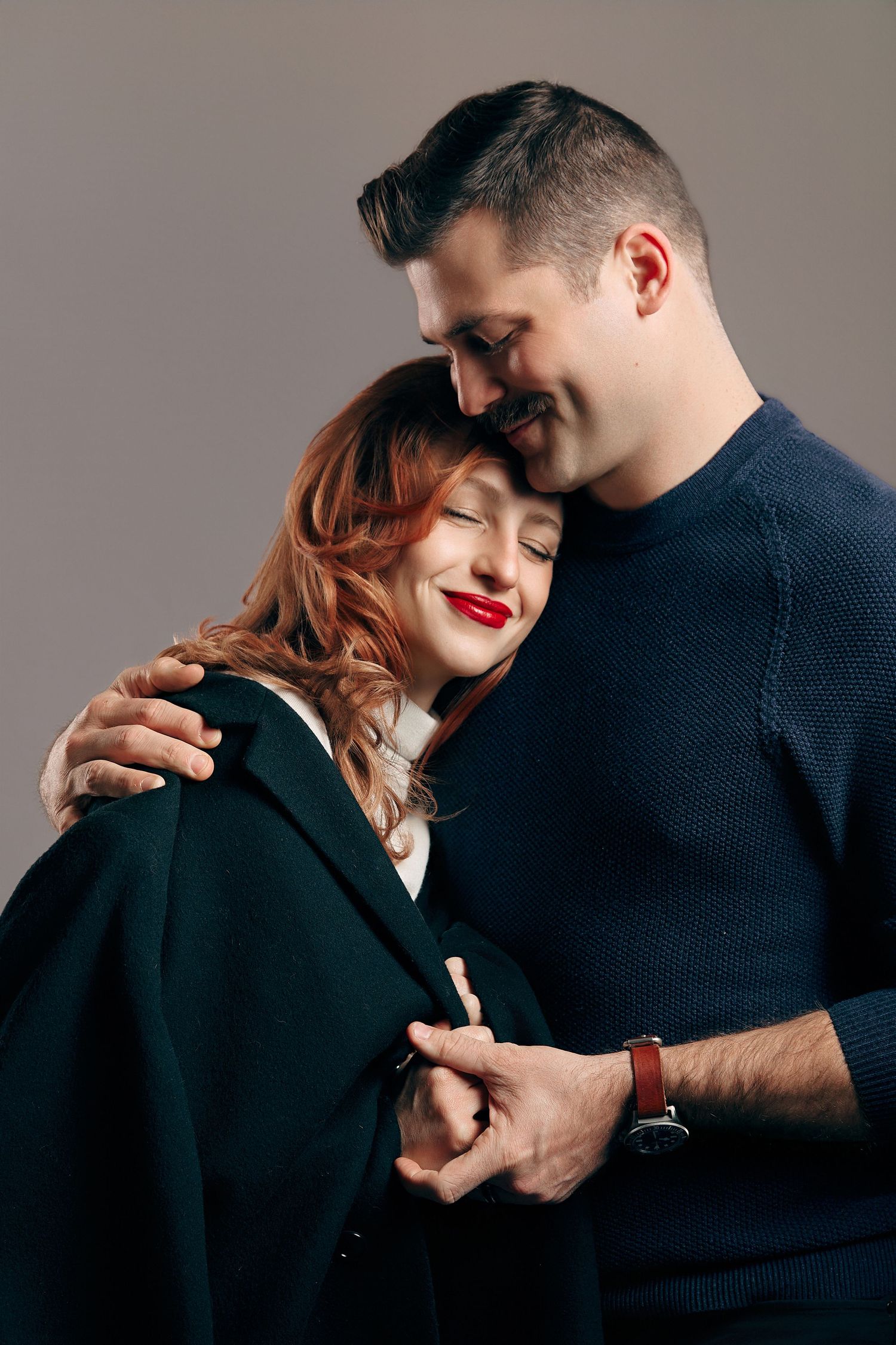Romantic couple embracing in studio, woman with red lips resting head on man's chest while he smiles warmly.
