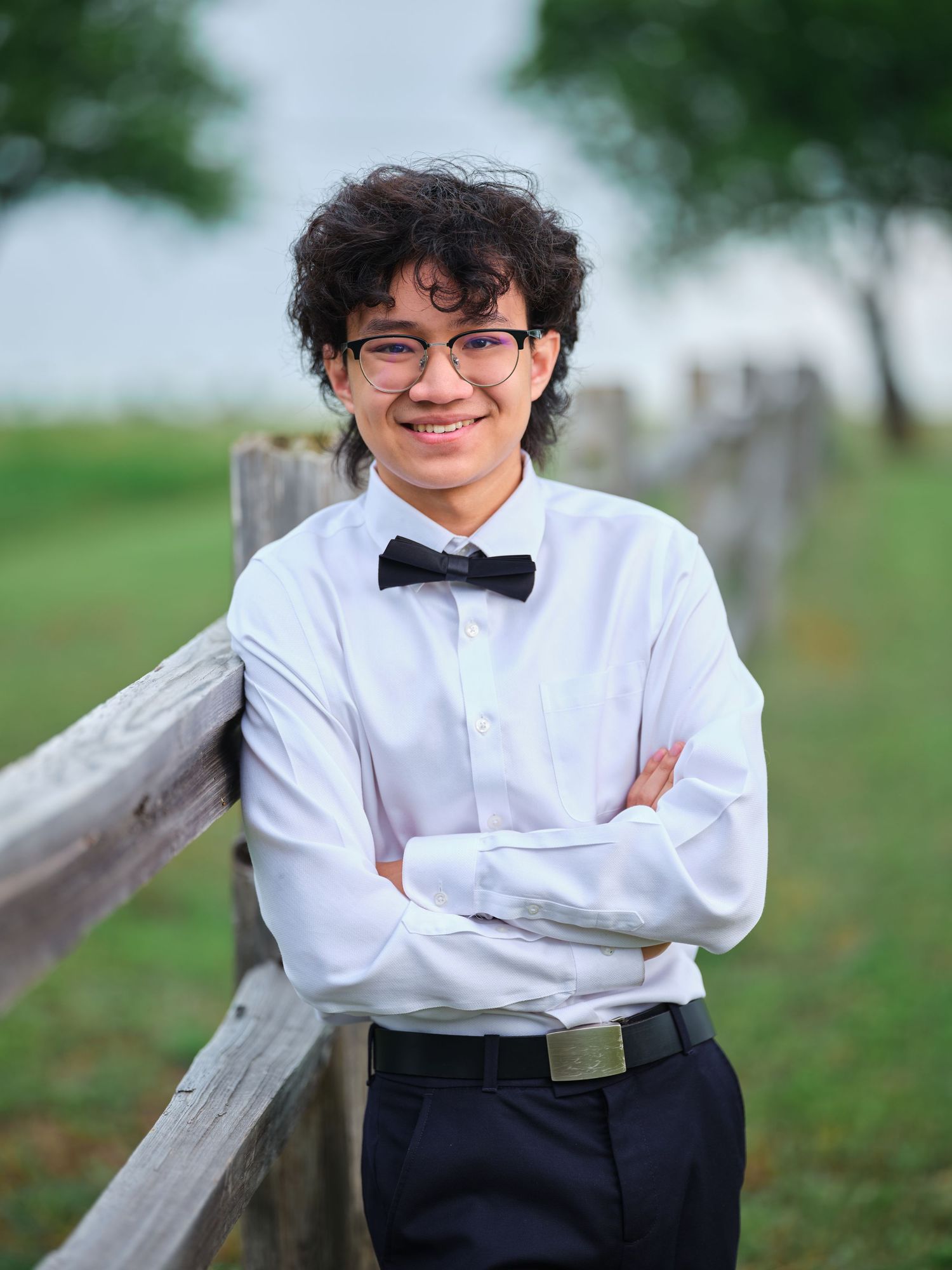 Smiling young man in white dress shirt and bow tie posing with crossed arms by a wooden fence outdoors.