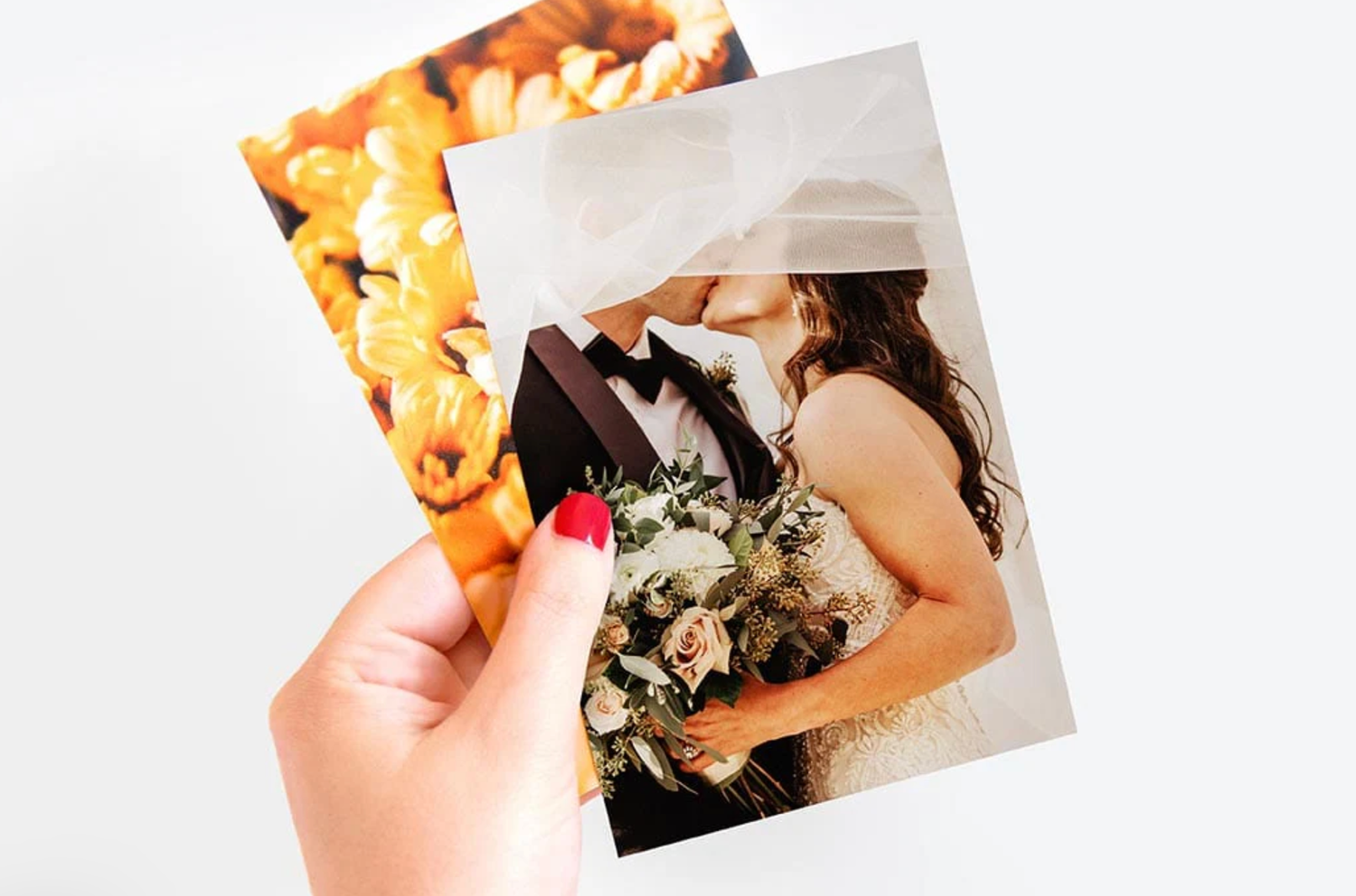 A hand with red nails holds two printed photos, the top one showing a wedding couple kissing under a veil.
