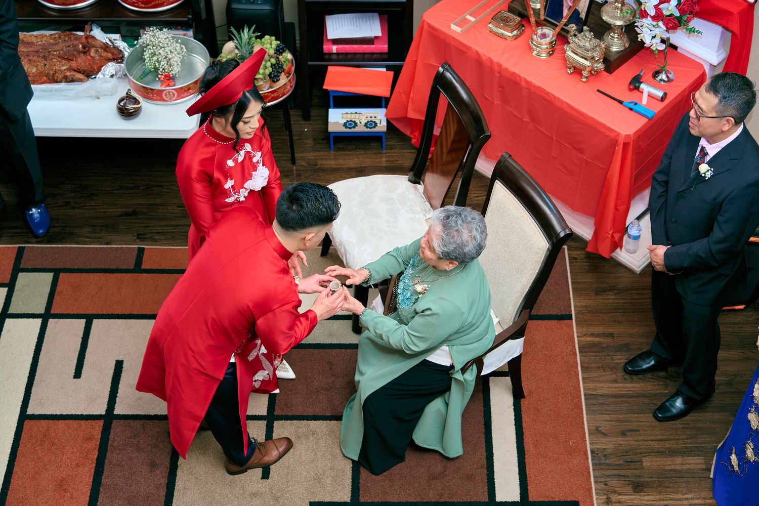 Traditional Vietnamese wedding ceremony with couple in red ao dai bowing to elderly relative indoors.