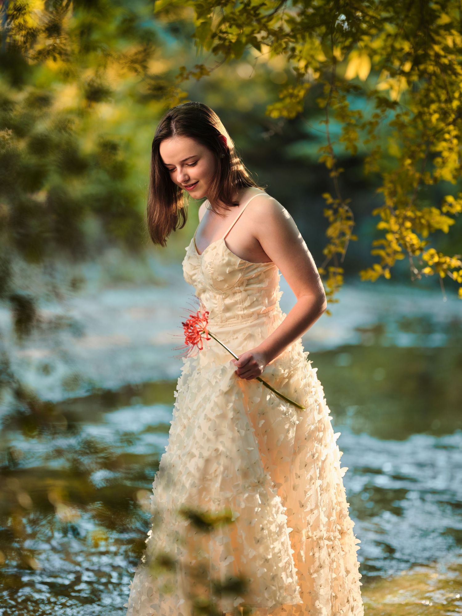 Elegant woman in ivory textured gown holding red flowers while standing in a serene reflective river.