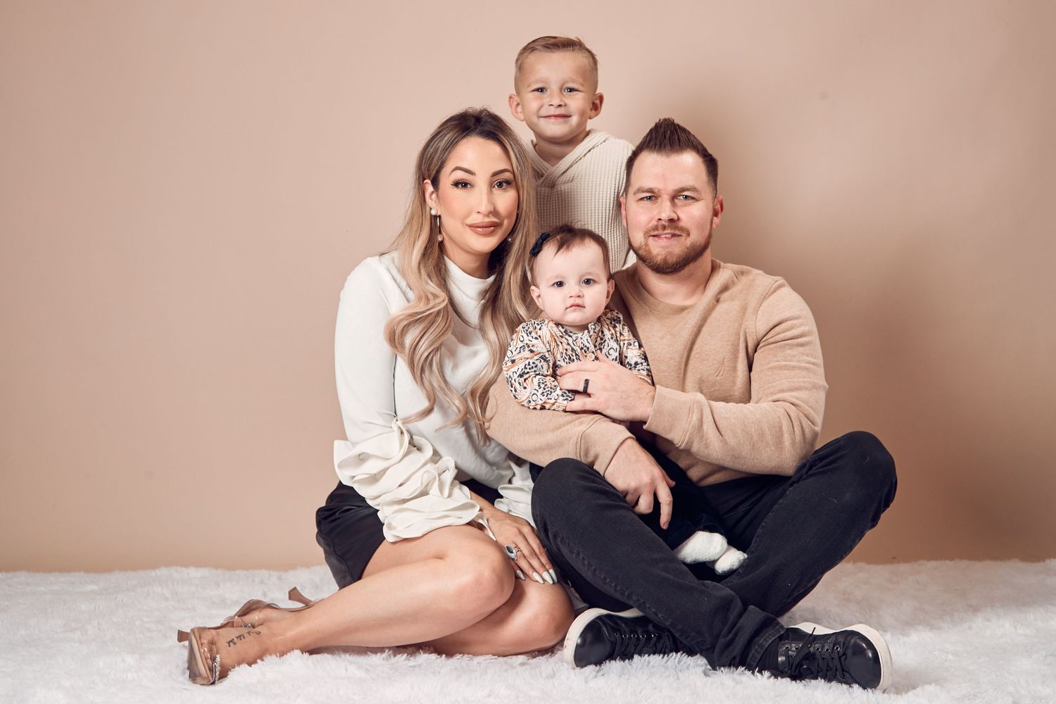 Young couple sitting on white fur rug with two small children in a warm-toned studio portrait.