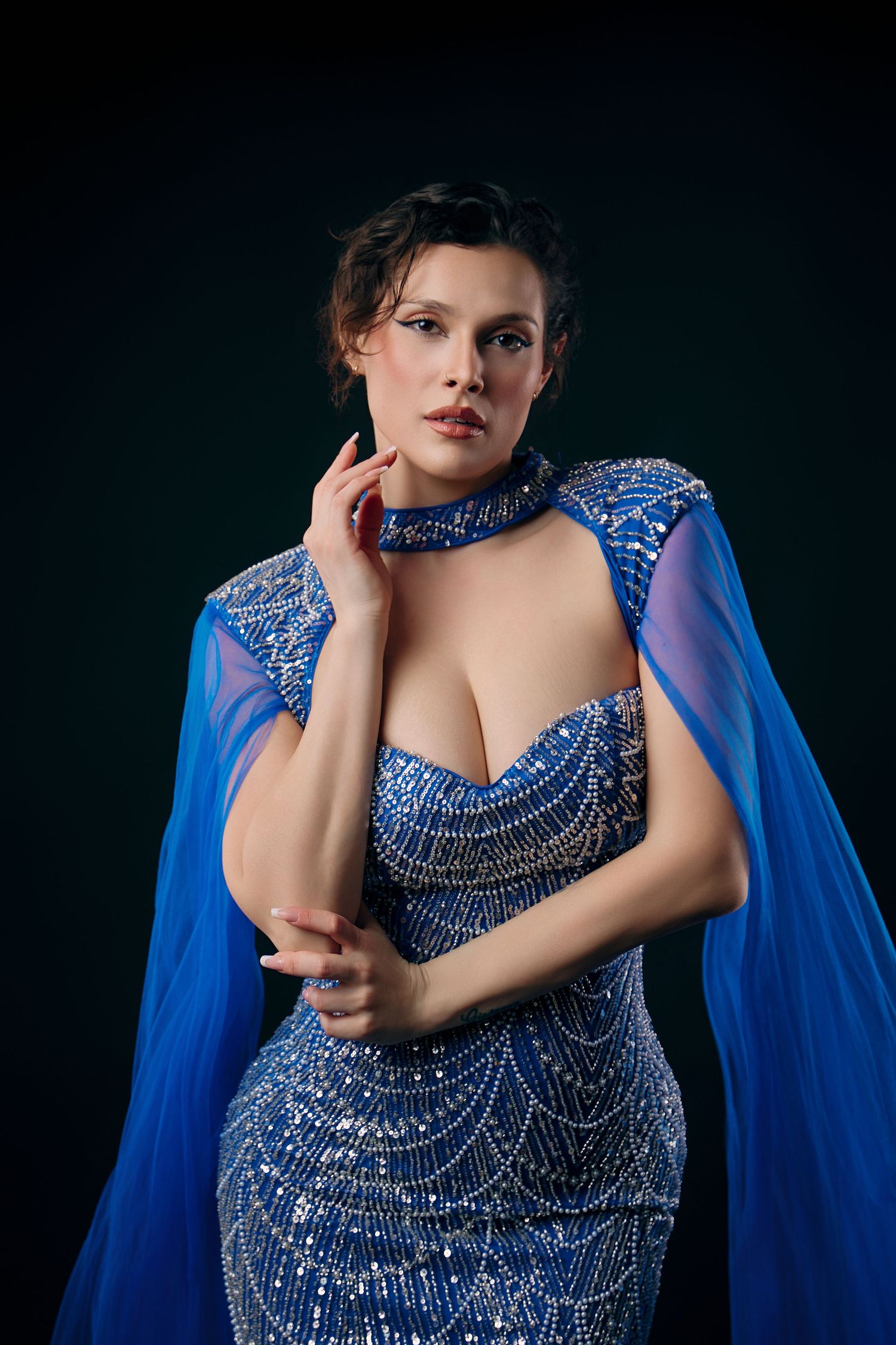 Elegant woman in sparkling blue sequin gown with cape sleeves poses against dark studio background.