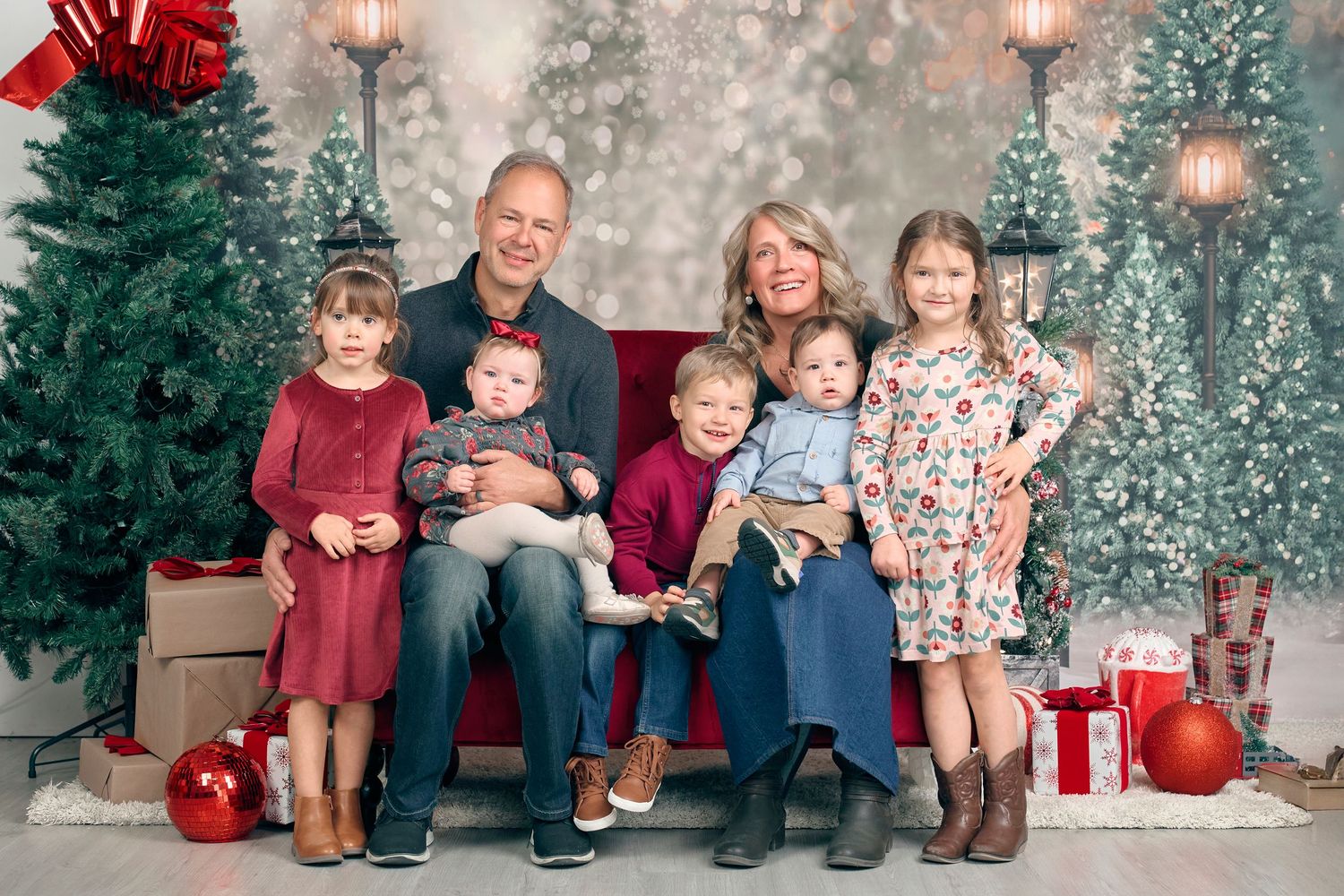 A family of seven with young children poses on a red bench surrounded by Christmas trees and gifts.