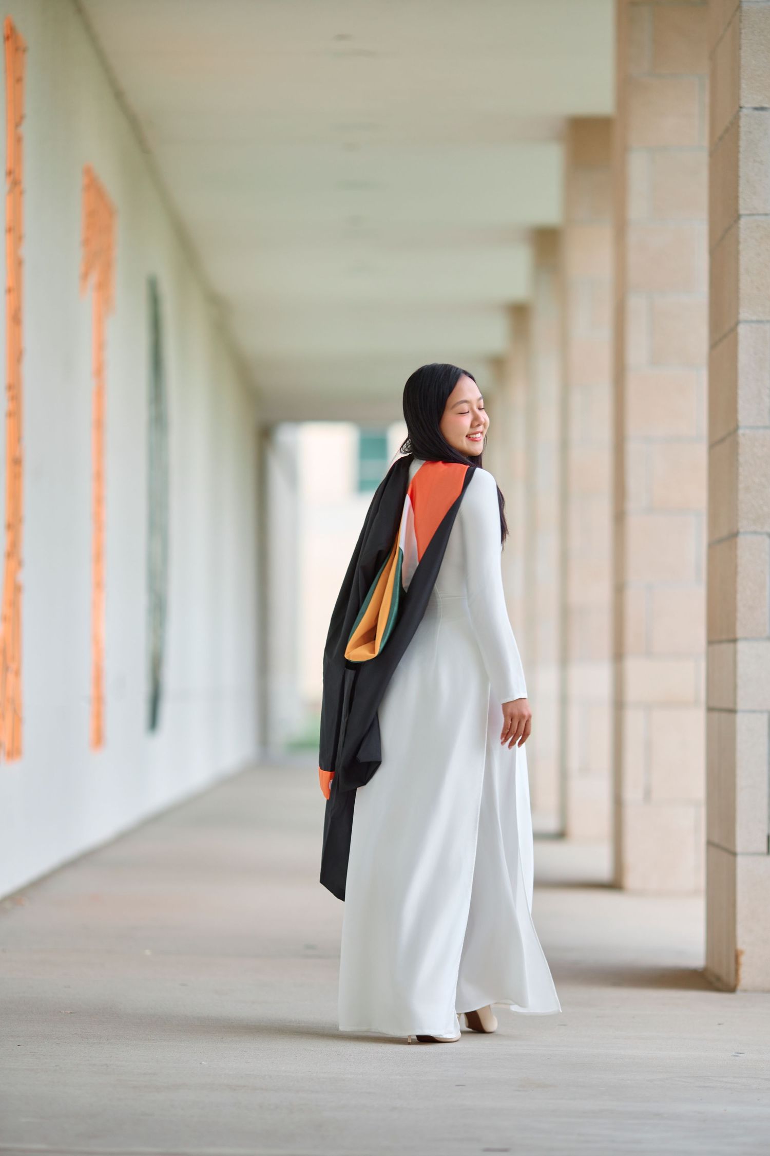 Female graduate in white dress with black and orange graduation regalia walking through corridor.