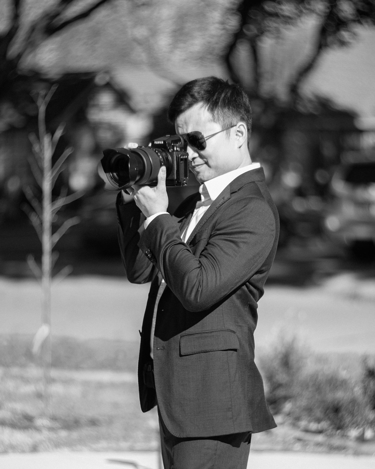 Man in suit and sunglasses photographing outdoors with a professional camera, black and white portrait.