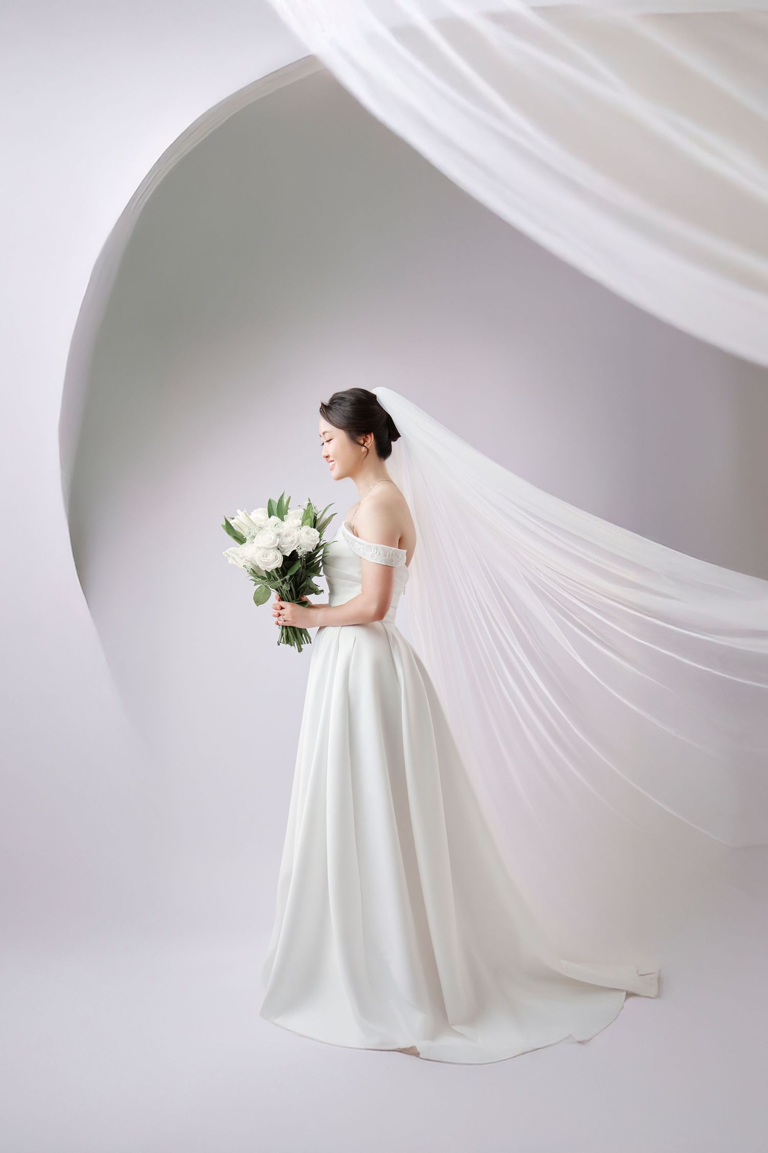 Graceful bride in off-shoulder white gown holding bouquet with flowing veil in studio.