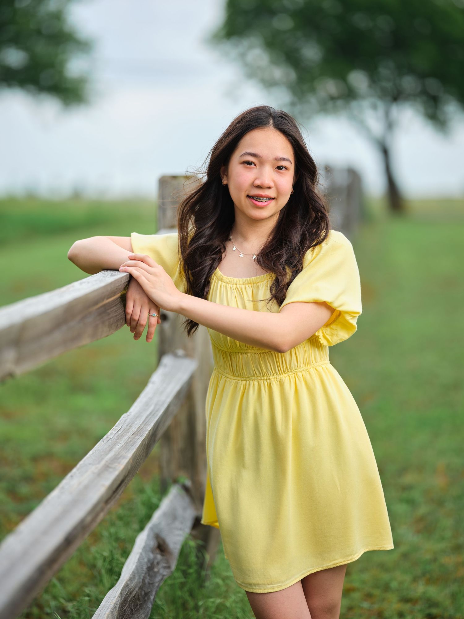 Smiling young Asian woman in yellow dress leaning on a rustic wooden fence in a green countryside setting.