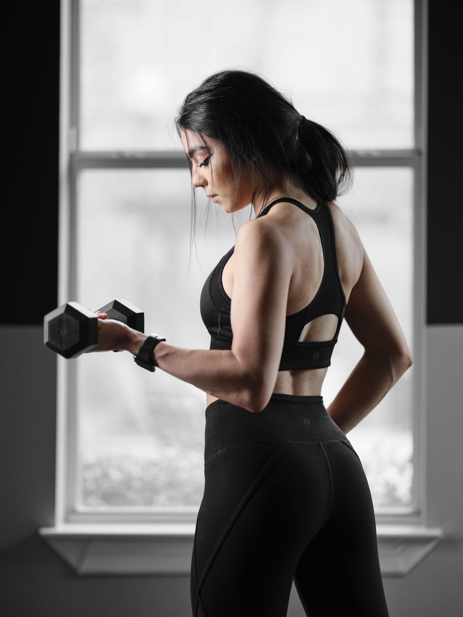 Athletic woman in black sports bra lifting dumbbell near window in gym setting.