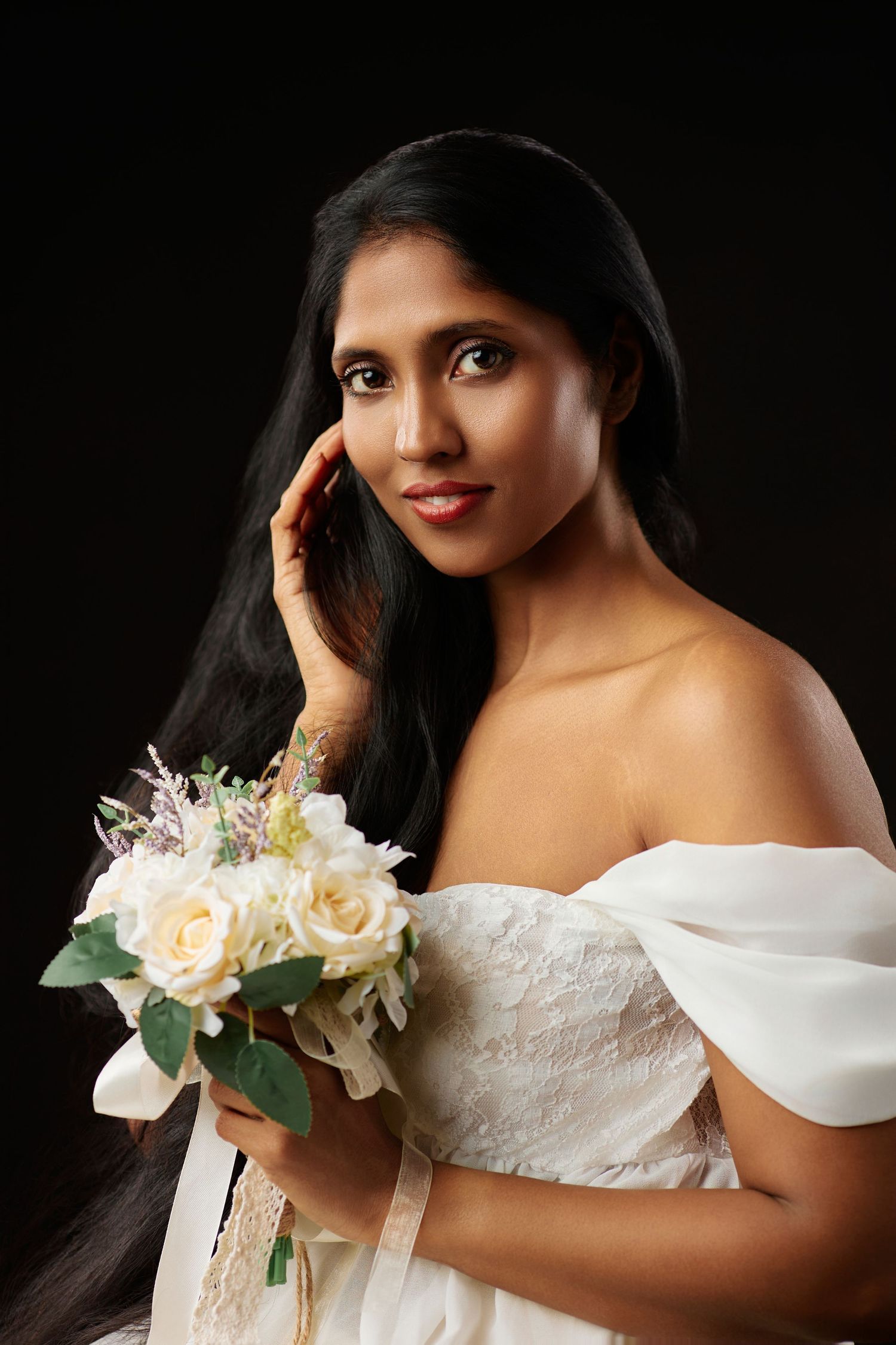 Beautiful South Asian bride in white off-shoulder gown holds white rose bouquet on dark background.