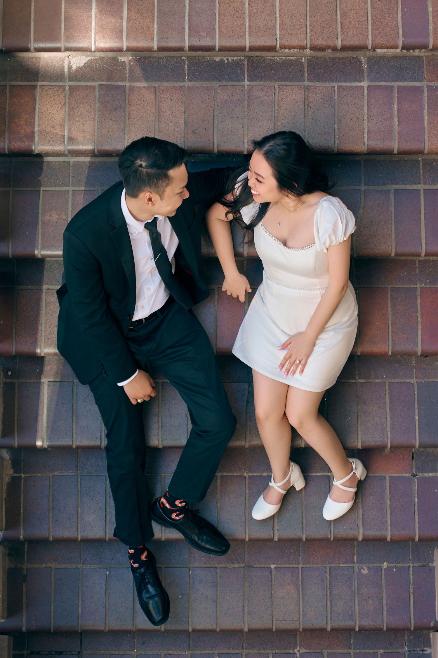 Aerial view of Asian couple in formal attire lying on tiled steps, smiling at each other.