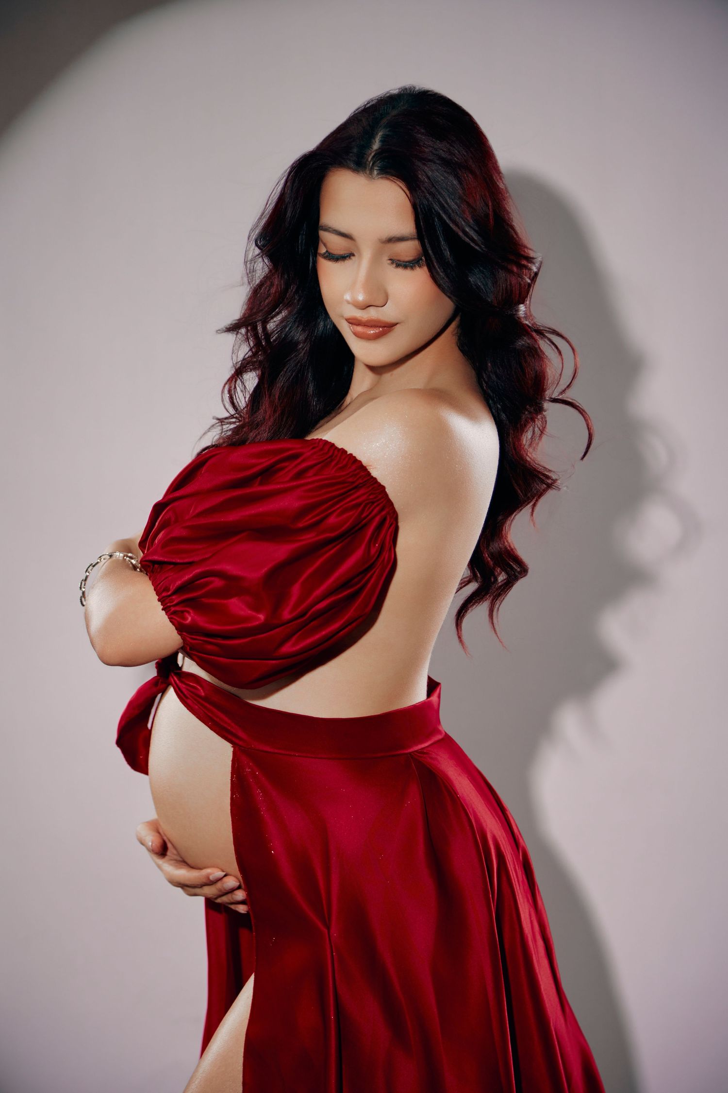 Elegant pregnant woman in a red satin off-shoulder gown holding her bare baby bump in a studio.