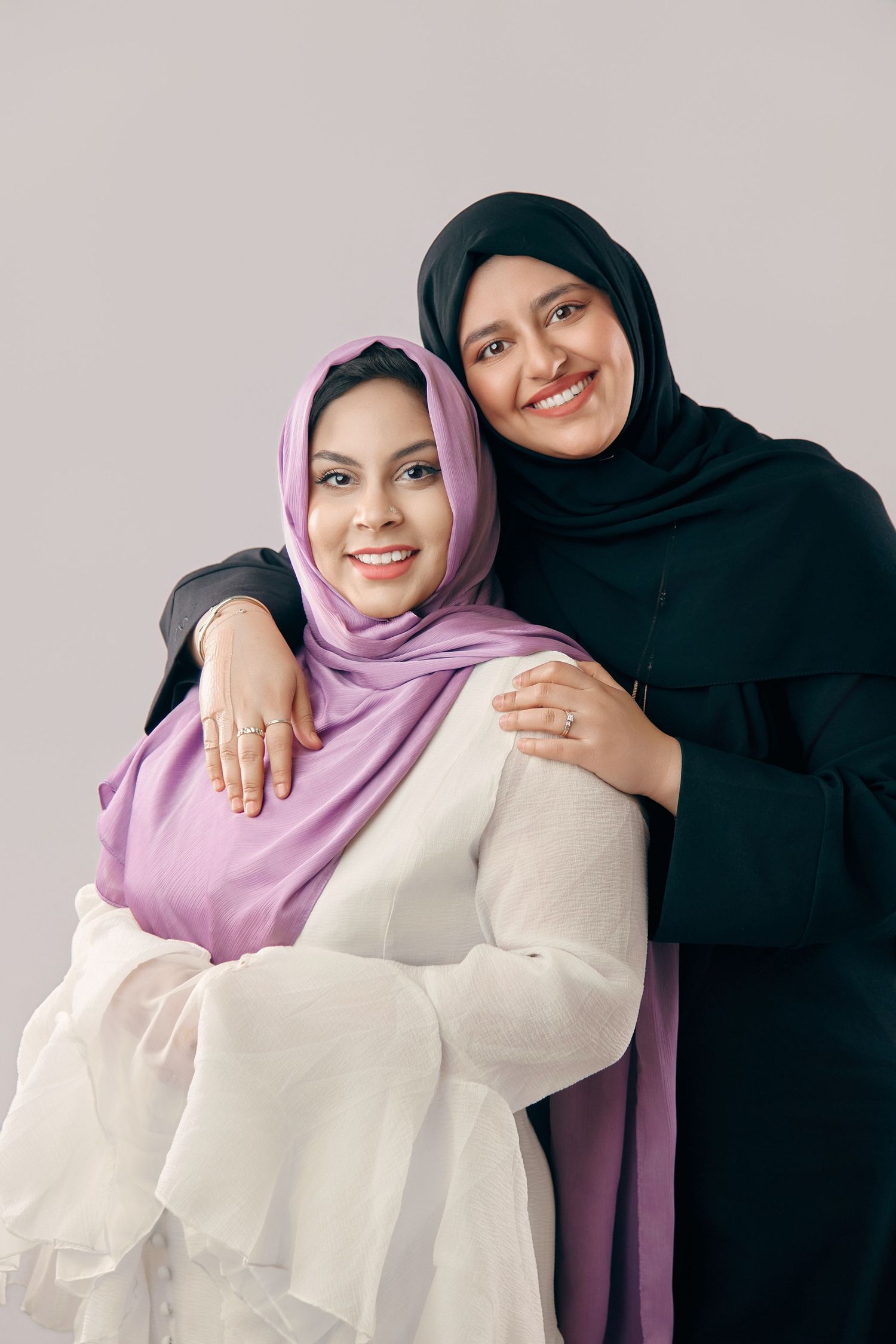 Two smiling Muslim women in hijabs embracing each other against a light gray background.