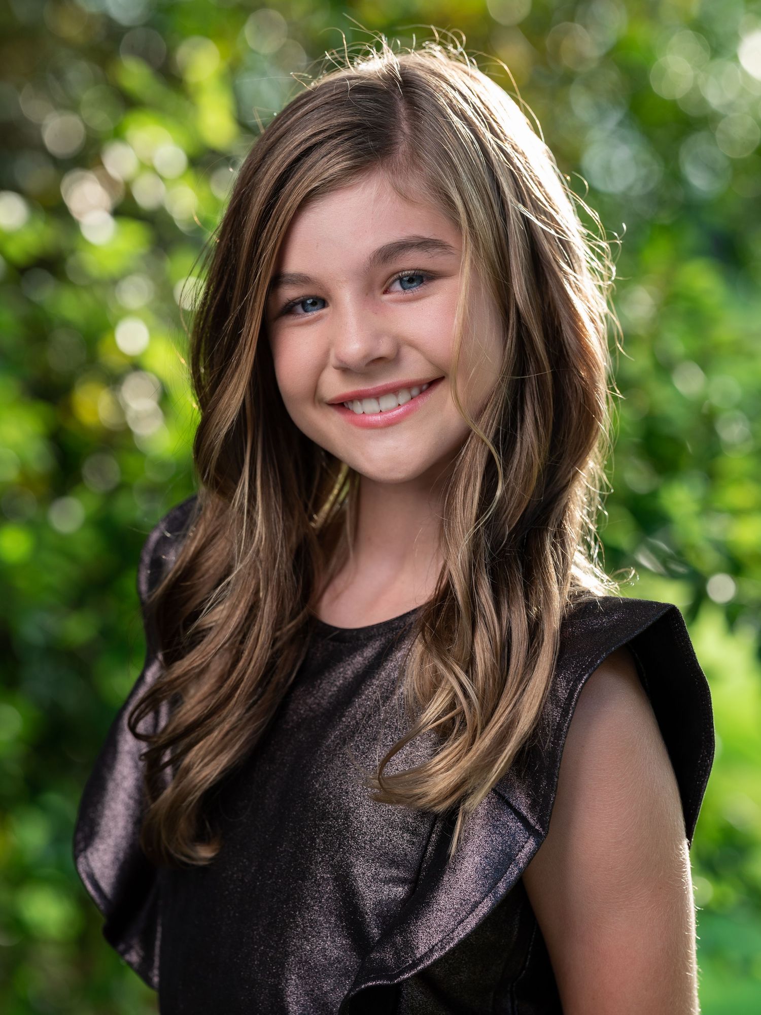 Smiling young girl with long brown hair wearing a black dress against a green bokeh background.
