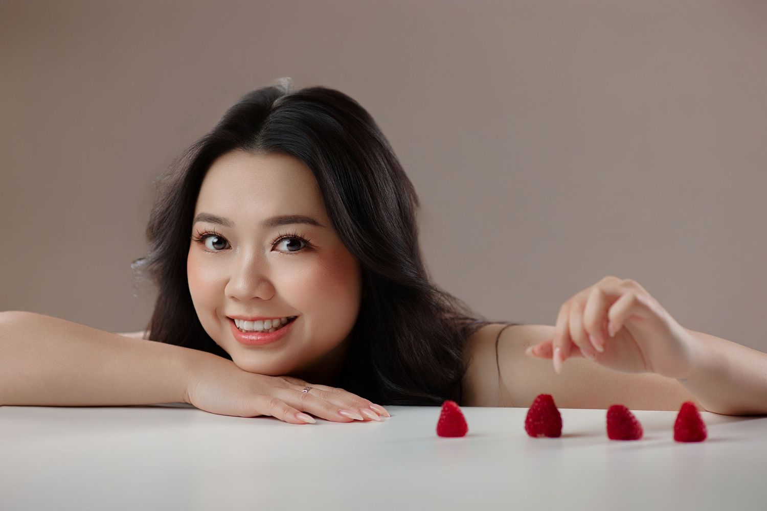 Smiling Asian woman lying on white table with raspberries in a beauty portrait.