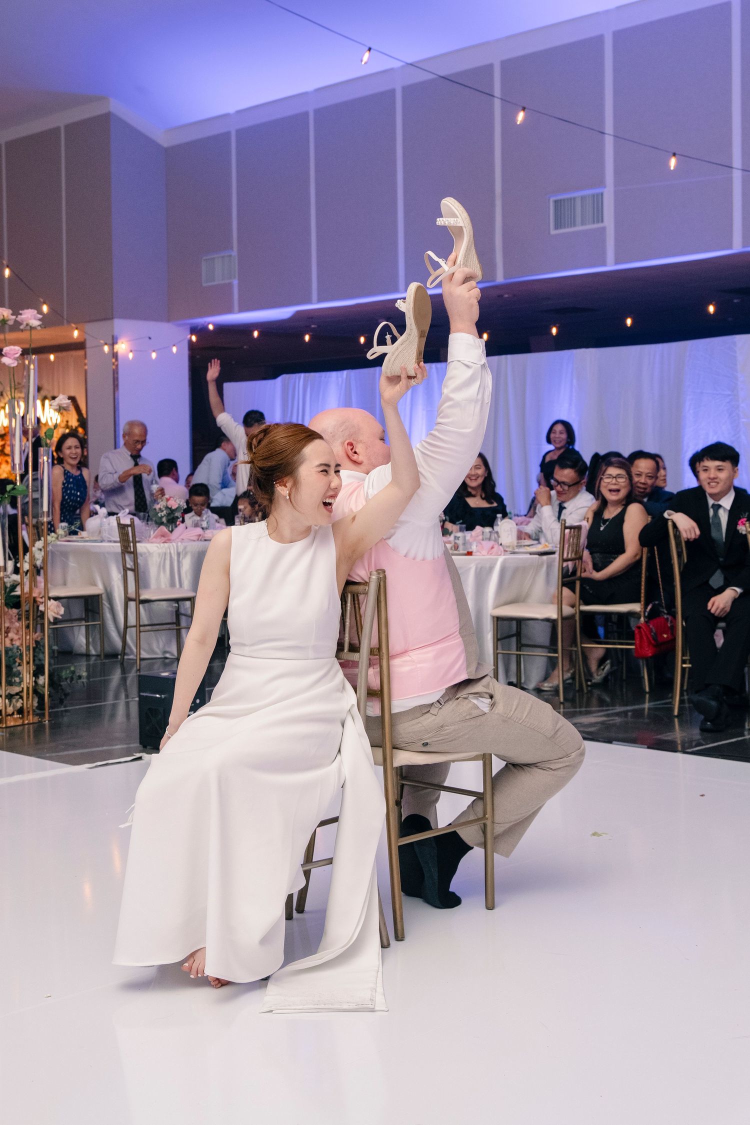 Groom playfully holds up bride's shoe during fun wedding reception game while bride laughs in chair.