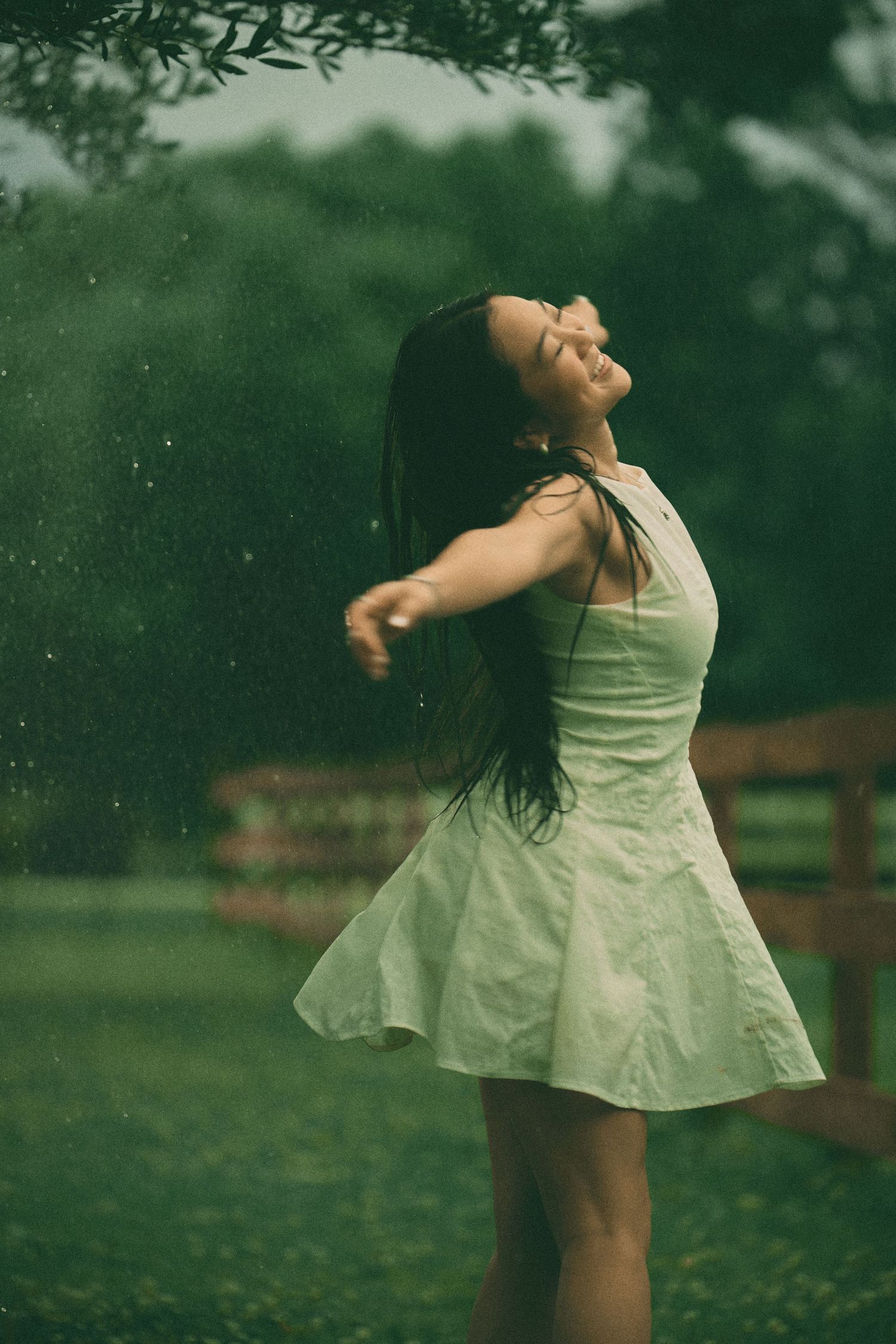 Young woman in white dress dancing joyfully in the rain outdoors, arms outstretched, eyes closed, surrounded by lush greenery.