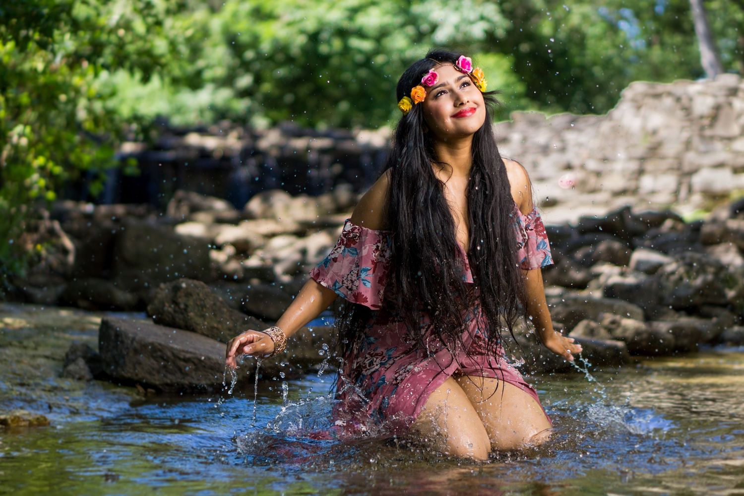 Joyful woman with flower crown splashing in a shallow rocky river wearing a floral dress.