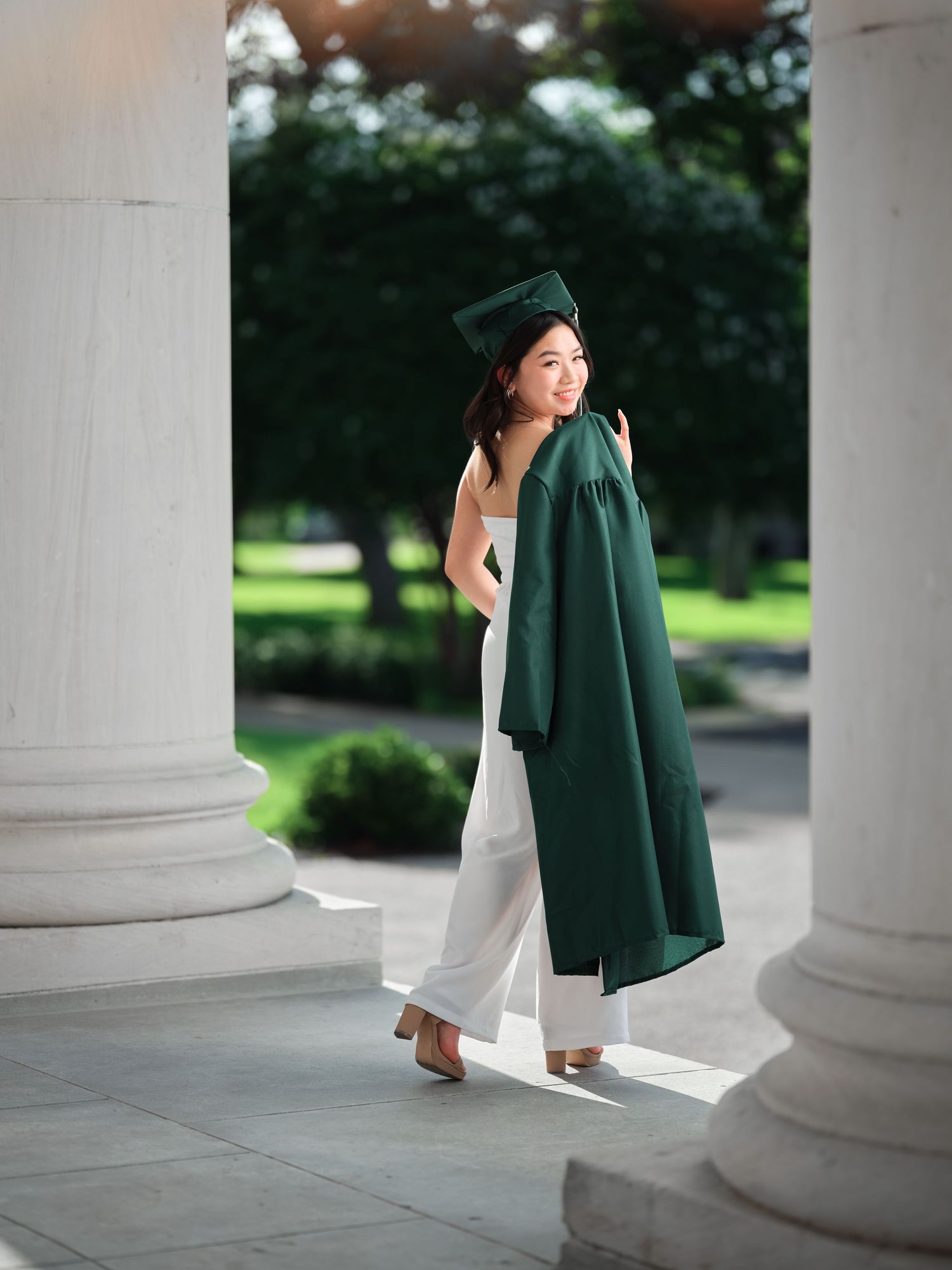 Young woman in green graduation cap and gown poses between white columns outdoors.