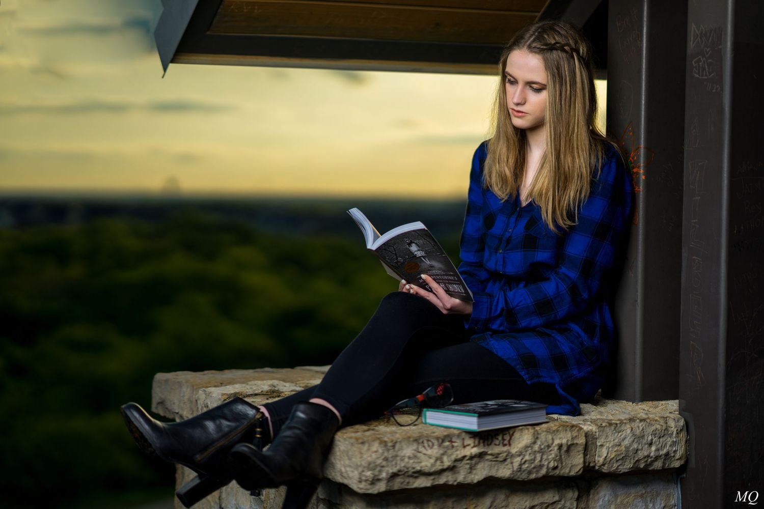 Teenage girl in blue plaid shirt reading book while sitting on stone ledge at sunset.