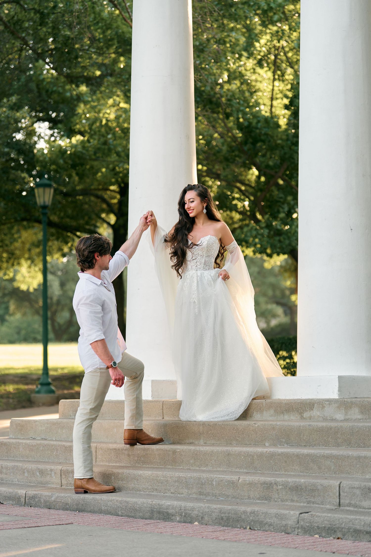 Bride in white gown holds groom's hand on steps between white columns in a sunlit park setting.