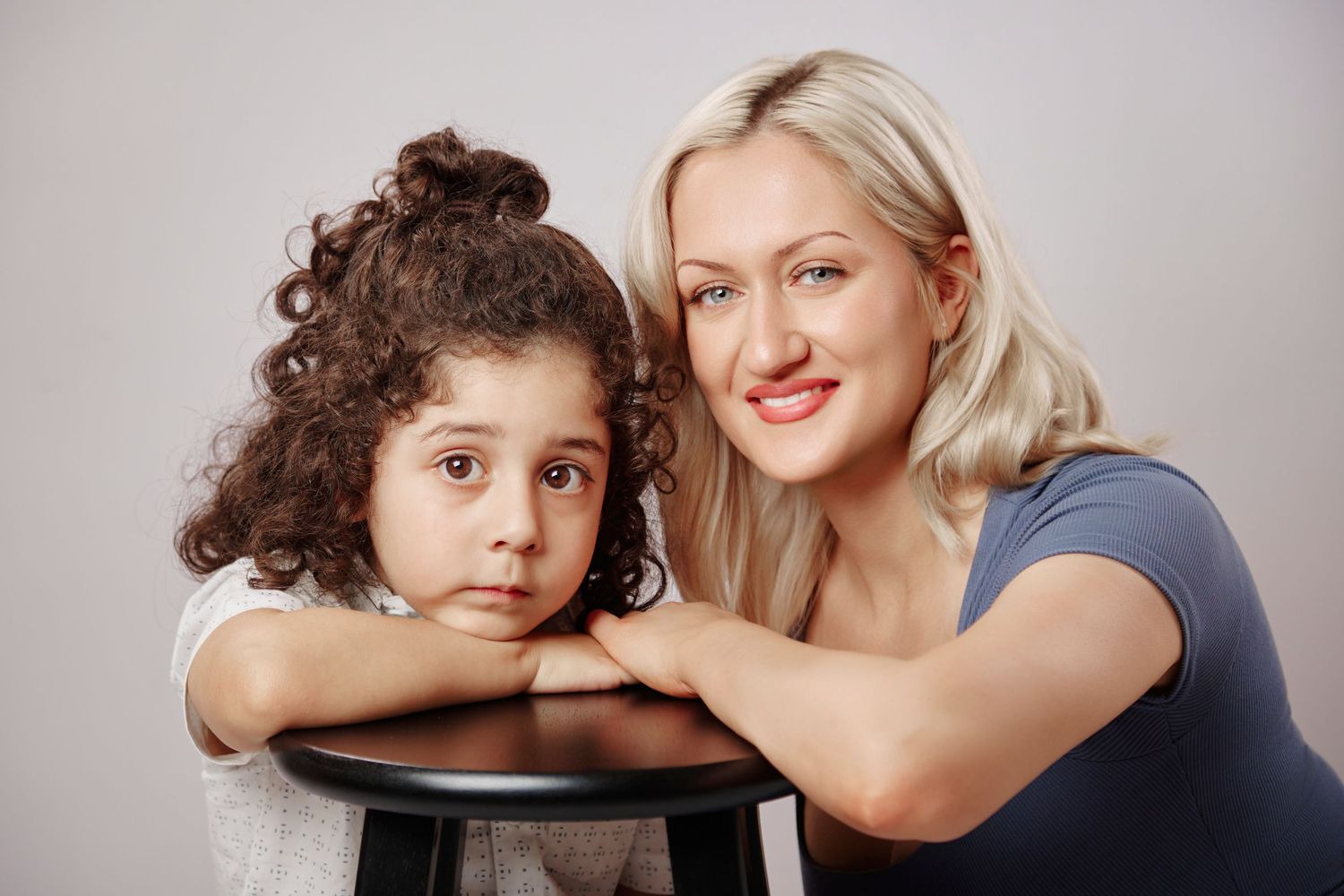 A blonde woman and a curly-haired young girl lean on a black stool together in a studio portrait.