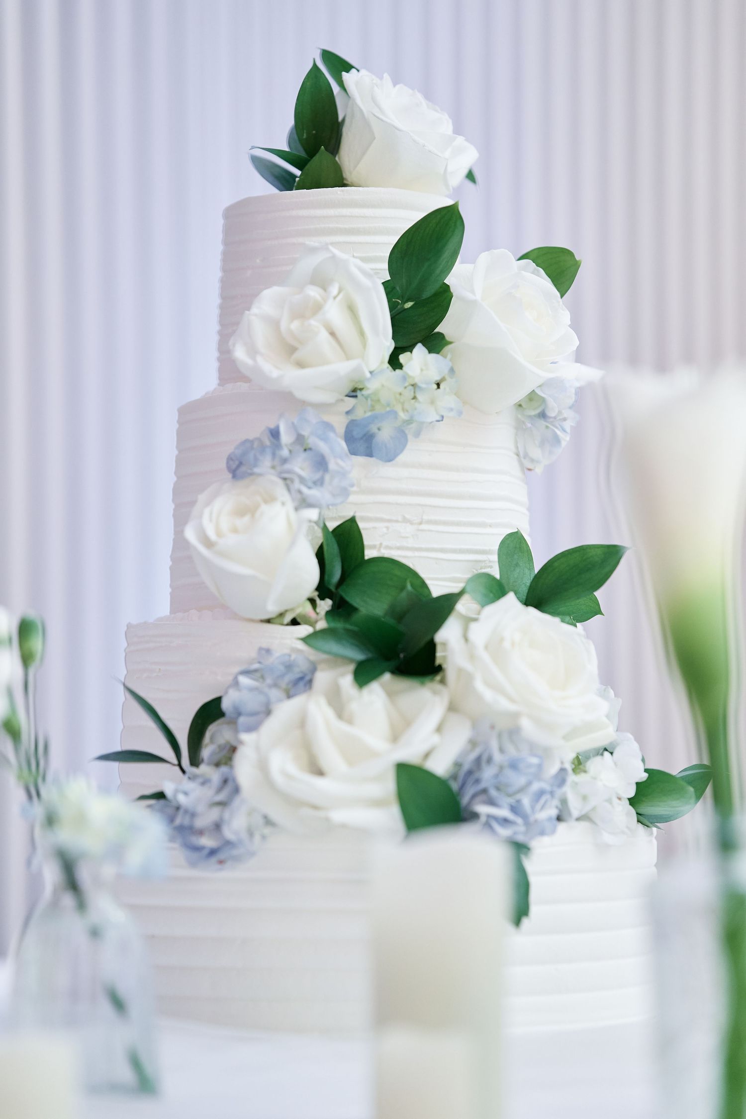 Elegant three-tier white wedding cake decorated with white roses and blue hydrangeas.