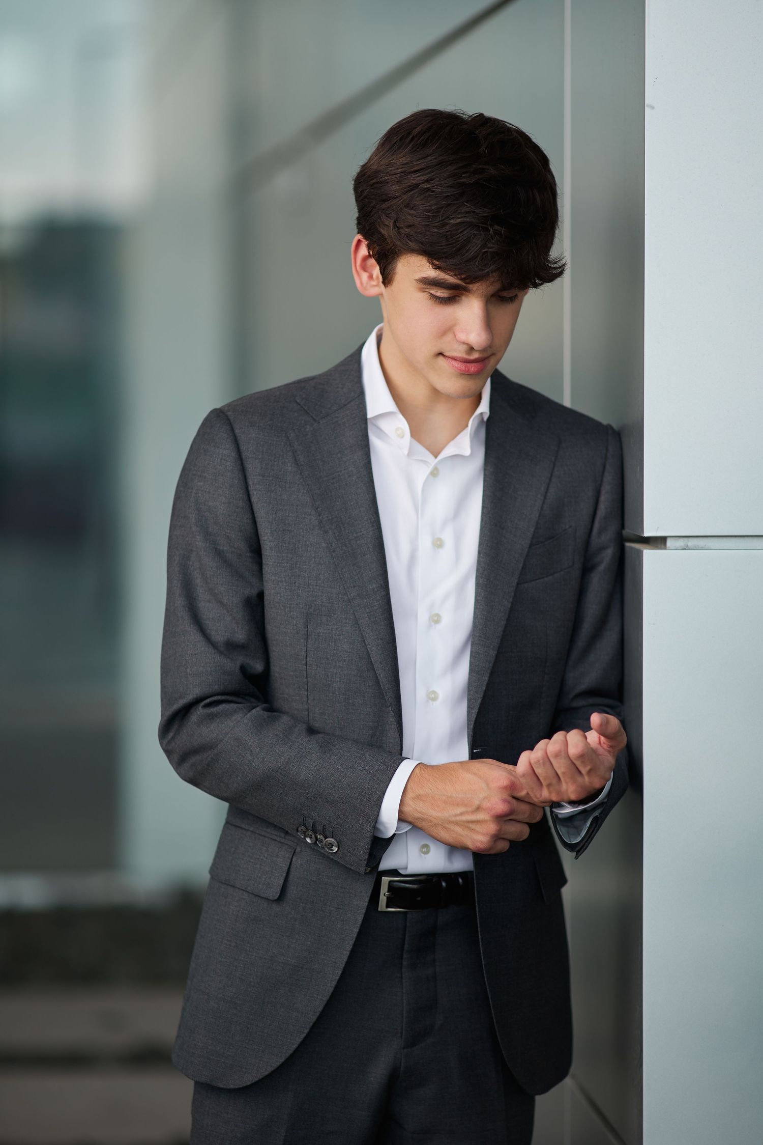 Young professional man in a gray suit adjusting his sleeve while leaning against a modern glass wall.