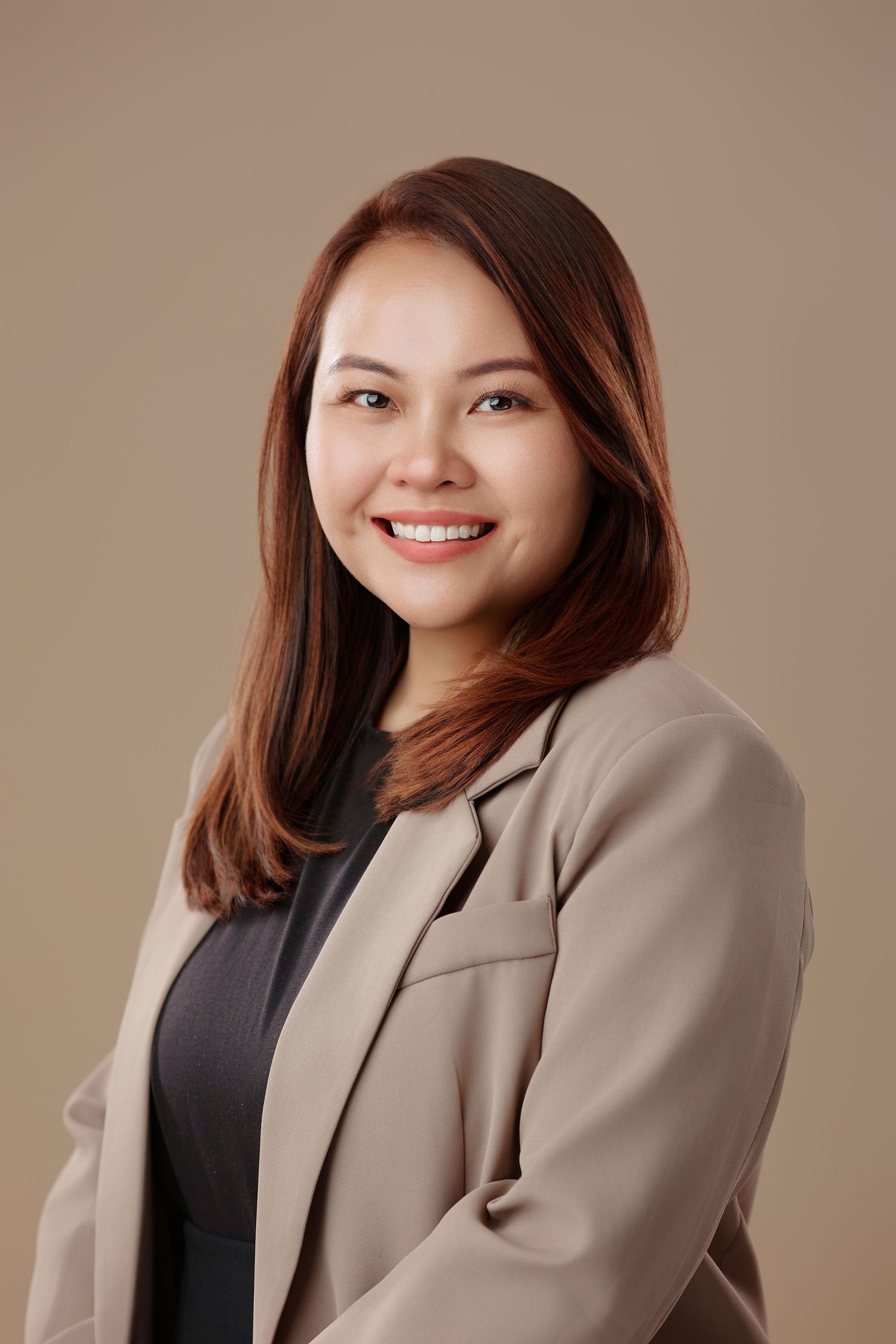 Professional portrait of a smiling Asian woman with brown hair wearing a beige blazer on tan background.