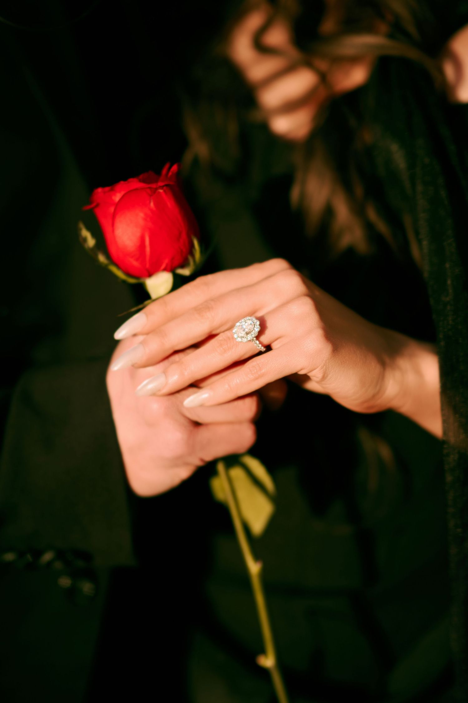 Close-up of a diamond engagement ring on a woman's hand holding a single red rose.