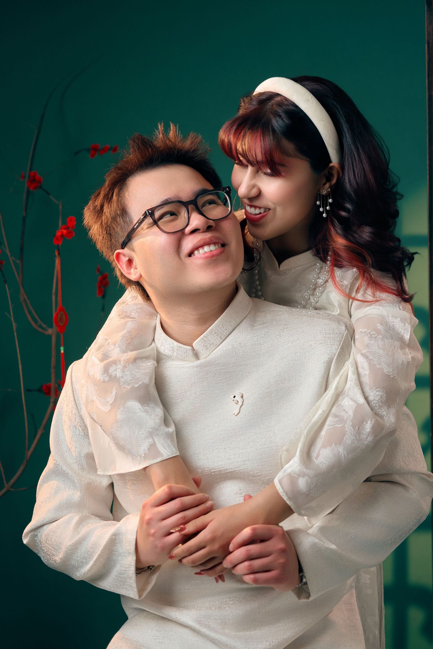 Couple in matching white traditional Vietnamese ao dai outfits embracing, smiling, against a green background.