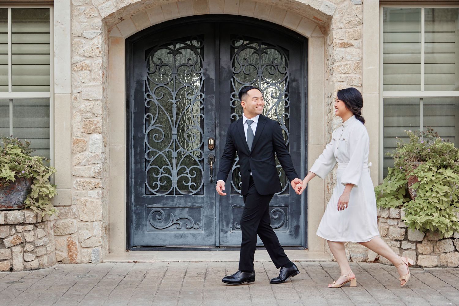 Couple holding hands walking in front of elegant stone home with ornate wrought iron double doors.