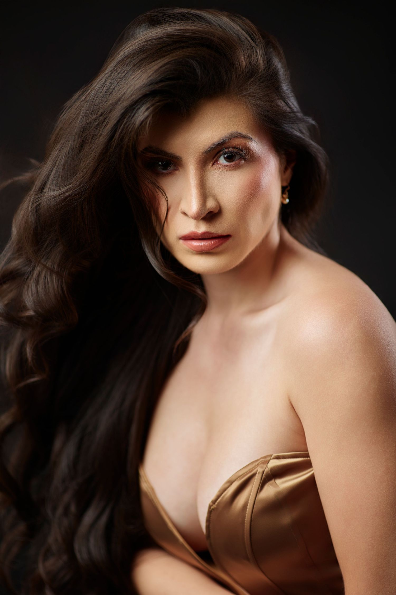 Glamorous woman with long dark wavy hair in gold strapless dress, dramatic studio portrait on dark background.