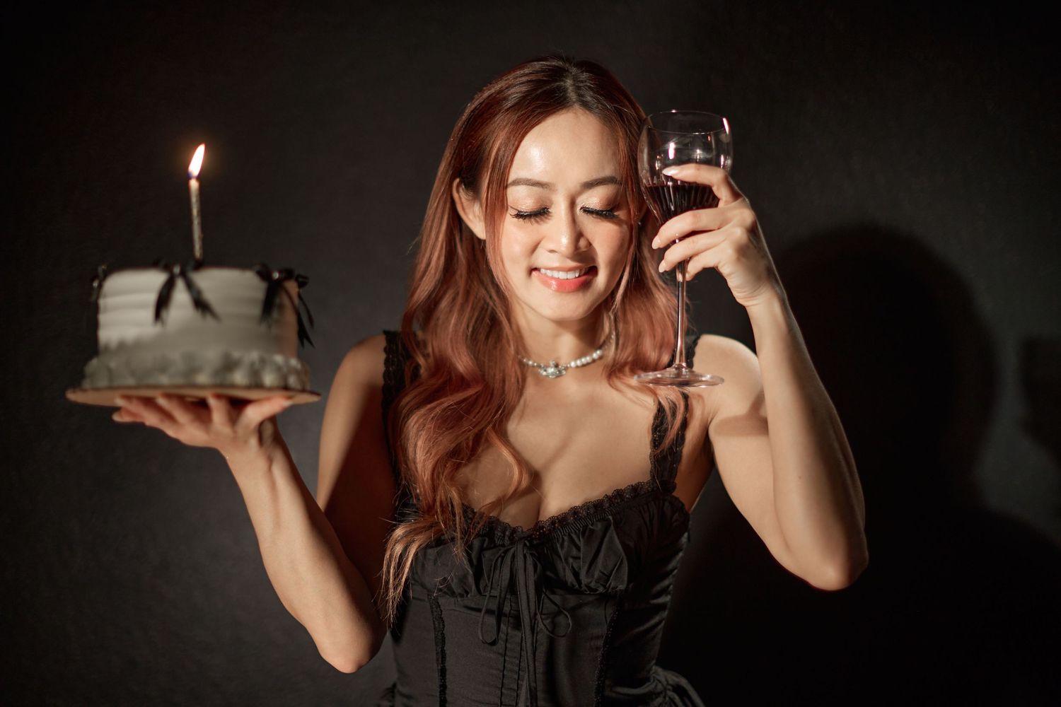 Smiling woman in black dress holding birthday cake and wine glass on dark background.