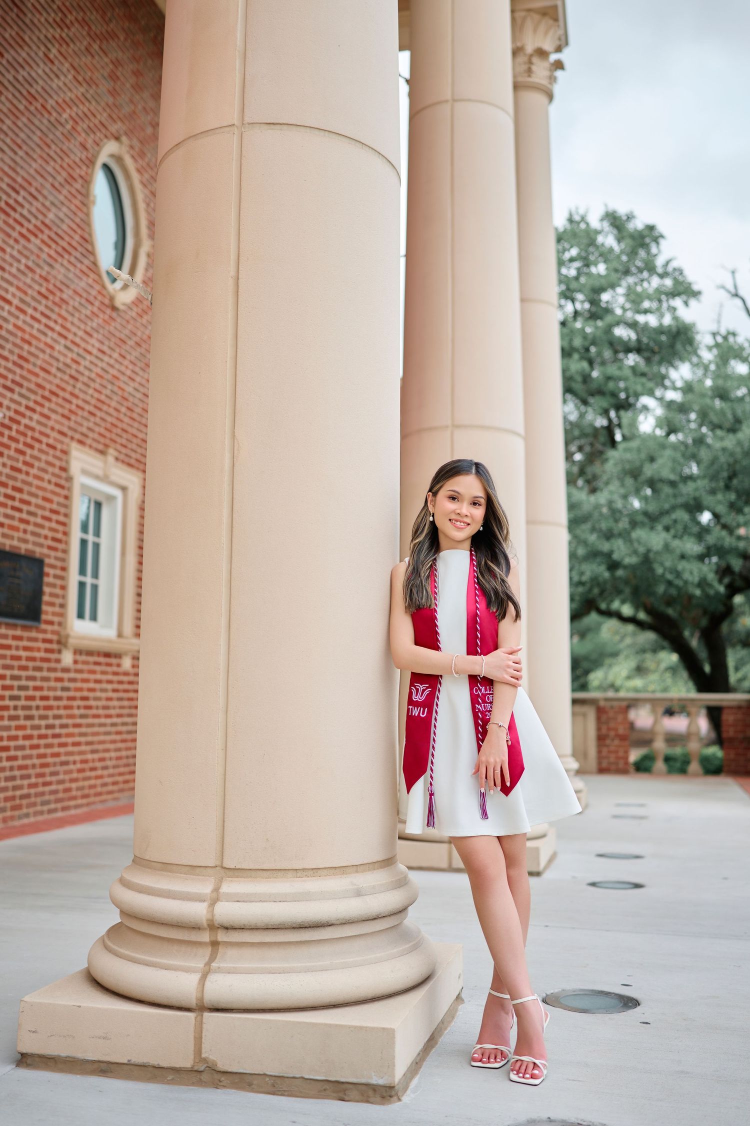 Smiling graduate in white dress with red stole poses between large columns.