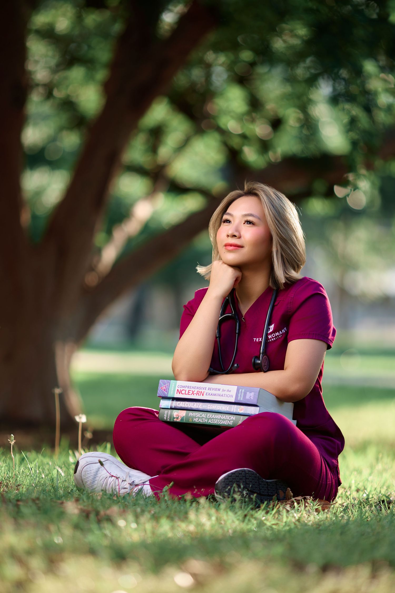 Nursing student in maroon scrubs sits under a tree with stacked textbooks.