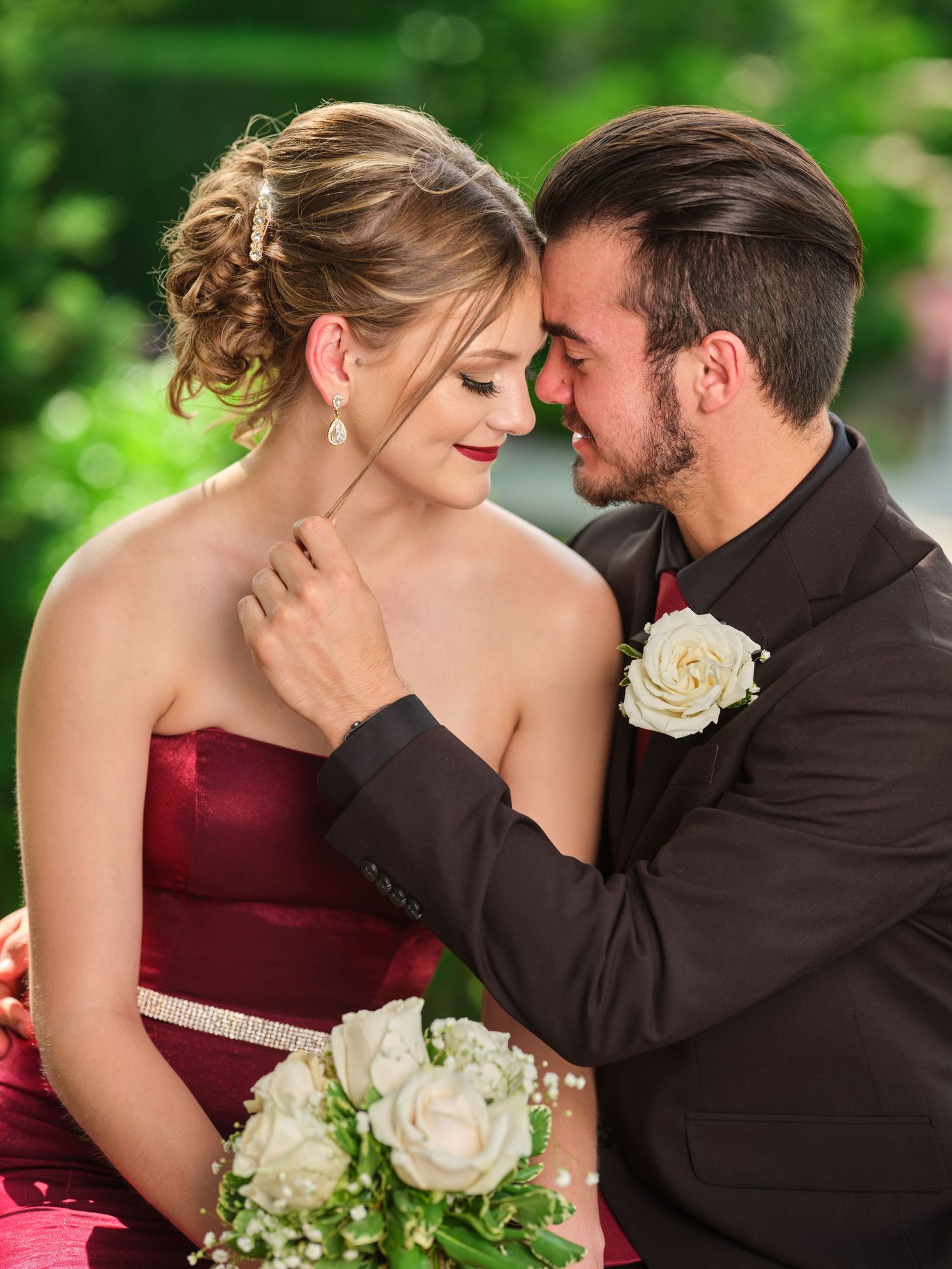 Young couple in formal wear sharing intimate moment, woman in burgundy gown holding white bouquet.
