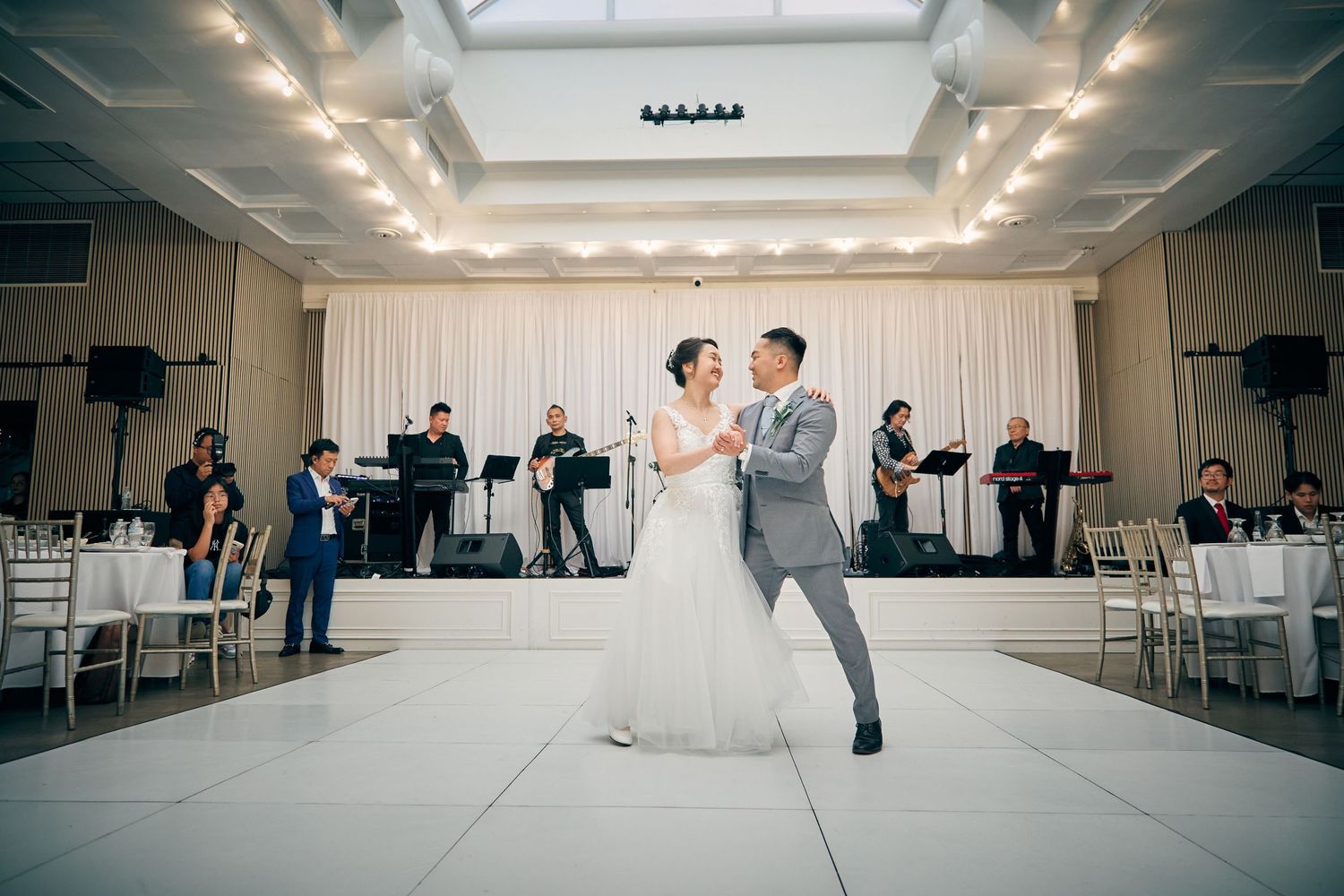 Bride and groom share their first dance on white dance floor with live band performing behind them.